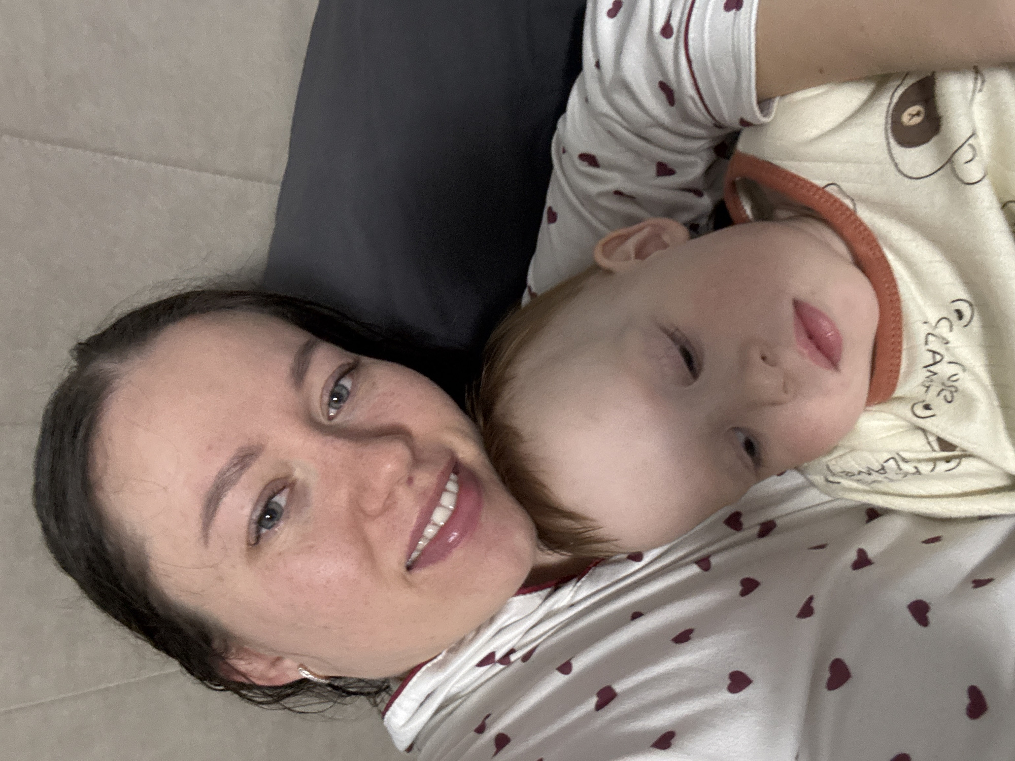A selfie shows a woman and a young child lying down together. The woman is smiling, with dark hair and blue eyes. The child is resting on the woman, with red hair and closed eyes. Both are wearing patterned pajamas. The background is a gray couch. The location is Ферма, Russia.
