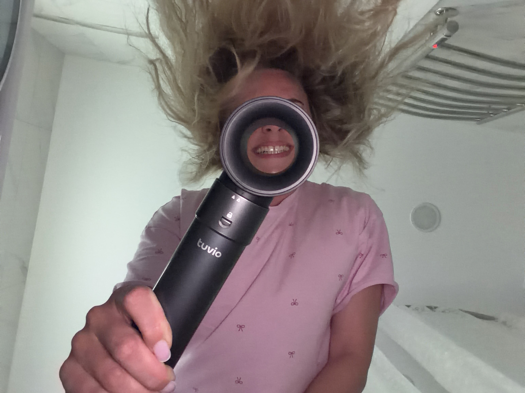 A woman with blonde hair stands in a bathroom holding a black "Tuvio" branded device in front of her face, partially obscuring her features, except for a glimpse of her teeth through the device's opening. She wears a pink t-shirt with a pattern of small bows. Her hair is styled in a voluminous, tousled manner, possibly indicative of being freshly dried or styled. The bathroom features a white wall and a towel warmer is visible in the background, along with a small ceiling speaker. The lighting is bright and even. The image's perspective is from a slightly low angle. The location in Votkinsk, Russia is not ascertainable from the image.