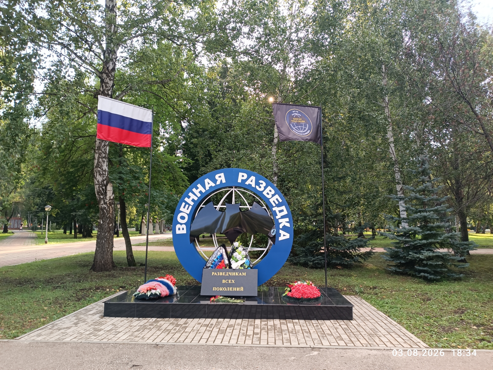 A monument with the inscription "военная разведка всех поколений" (military intelligence of all generations) is located in a park in Sterlitamak, Russia. The monument is a blue circle with a globe inside, and a bat silhouette. Wreaths of flowers are placed at the base of the monument. A Russian flag and a black flag with a circular emblem are visible behind the monument. The environment is a park with trees, grass, and paved walkways. The date "03.08.2026 18:34" is visible in the bottom right corner of the image, indicating a possible timestamp, though it appears to be a future date. There are no people clearly visible in the immediate vicinity of the monument.