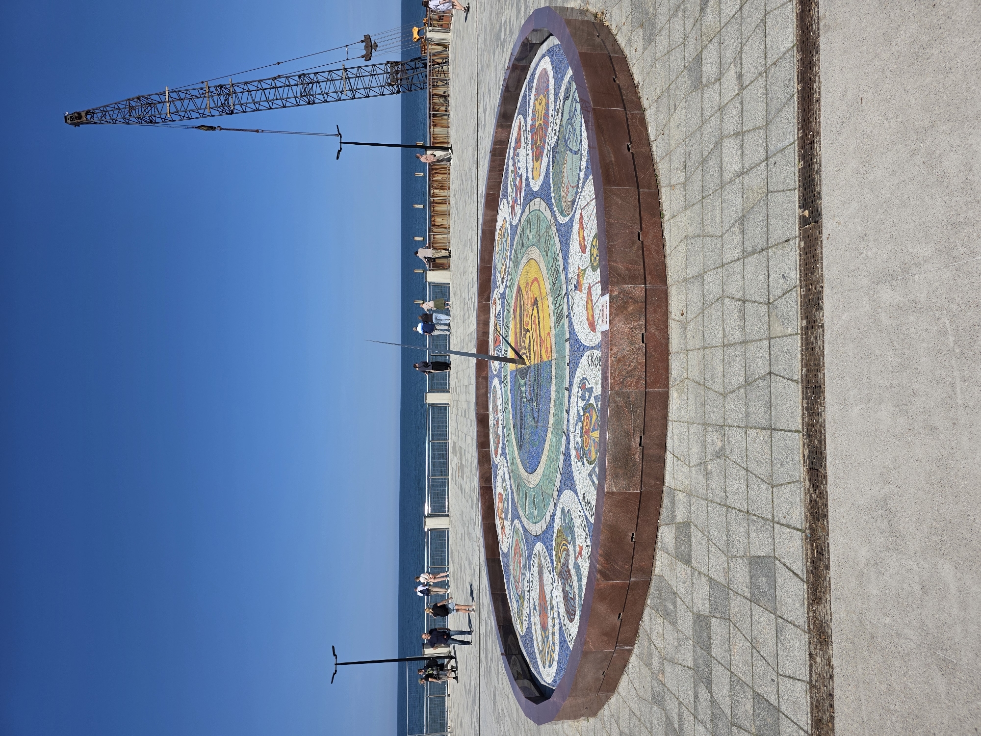 In the city of Svetlogorsk, Russia, a large, circular sundial is the central focus of this image, positioned on a paved plaza near the sea. The sundial features a mosaic surface with intricate designs, divided into sections, and a prominent gnomon, indicating the time. Surrounding the sundial is a raised copper-colored border.
In the background, the deep blue sea stretches to the horizon under a clear, bright blue sky. A promenade, lined with a metal railing, separates the plaza from the beach. Several people are visible on the promenade, some standing and observing the view, others walking.
To the right side of the frame, a large construction crane with a prominent boom stands tall against the sky, suggesting ongoing development or maintenance work. The ground in the foreground is paved with large, square tiles in a diamond pattern, with a metal grate running along the edge where it meets a concrete path. The overall scene depicts a public space on a sunny day, with elements of leisure, art, and construction.
