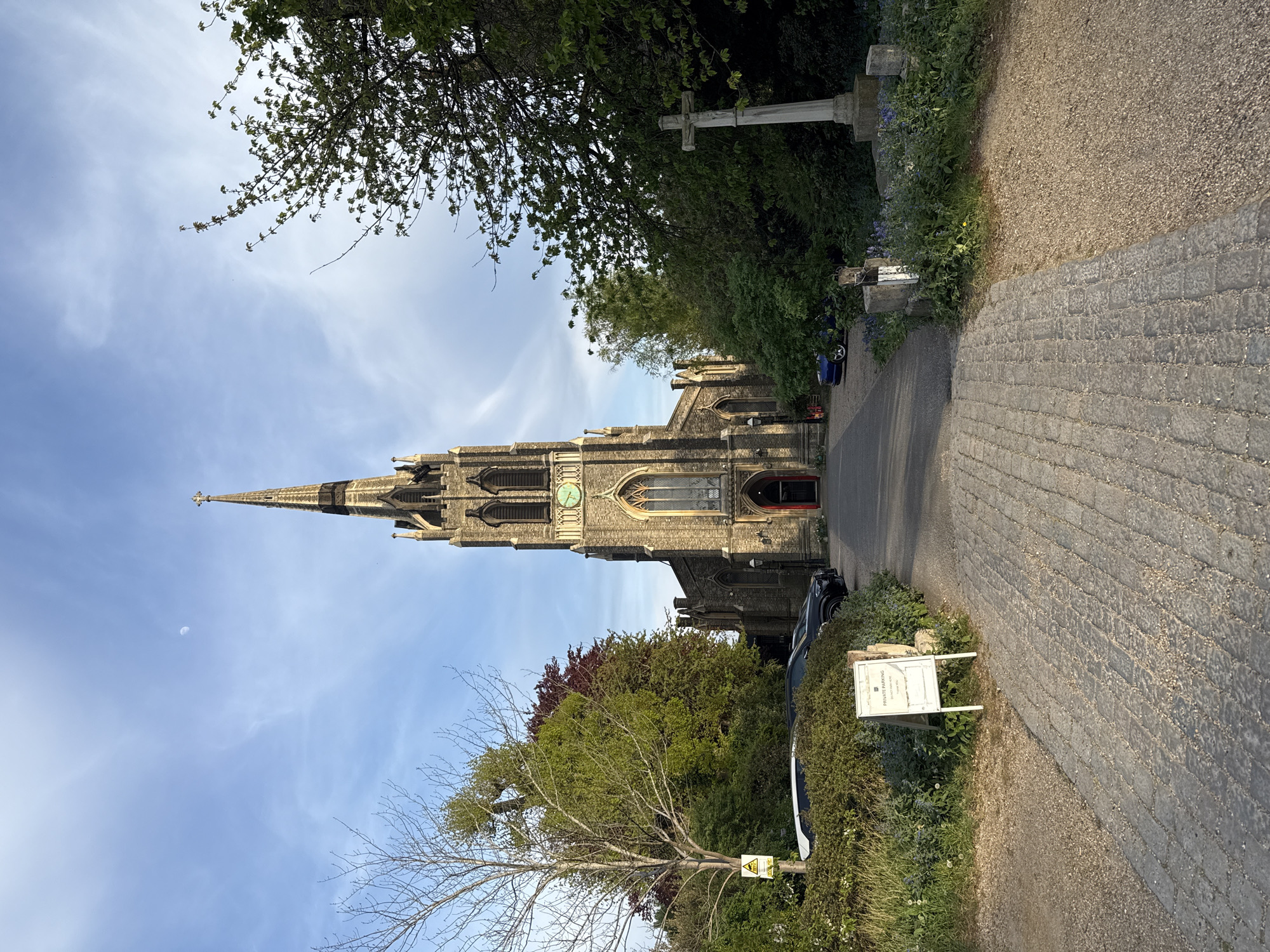 A church with a tall spire dominates the frame. The sky behind the church is blue with some clouds. A stone path leads towards the church. The church is surrounded by greenery, including trees and bushes. There's a sign on the right side of the path, and a stone cross is visible further down the path. A vehicle is parked near the church. The overall location appears to be rural.
The photograph is taken at St. Peter's Church in Seale, Surrey, England.