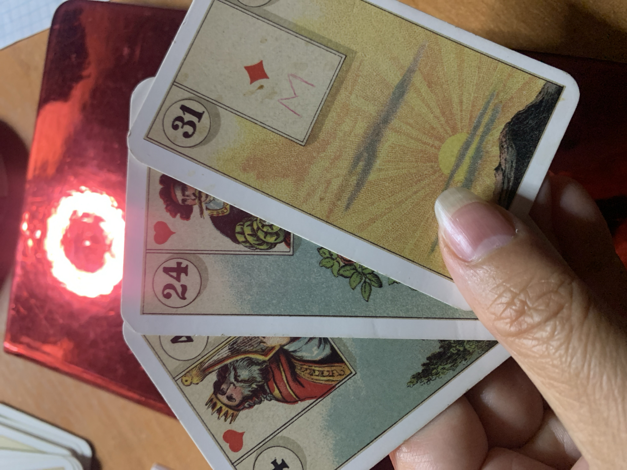 Three playing cards are fanned out in a person's hand. The leftmost card features a king with a crown and beard, playing a lyre, with a heart symbol above. The middle card depicts a man with a hat and mustache, and the number "24" in a circle. The rightmost card shows a sunset over mountains and the number "31" in a circle, with a red diamond symbol and a pink "M" drawn on it. The background includes a red, reflective surface with a bright, circular light. The image is cropped to focus on the cards and hand, and does not depict any specific location in Sevastopol, Ukraine.