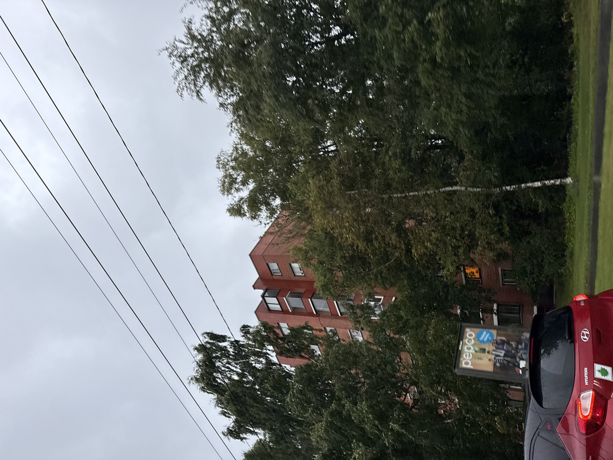 The image depicts a view of a red brick apartment building with multiple windows, partially obscured by trees. Overhead power lines are visible against a cloudy sky. In the foreground, the rear of a red Hyundai car is visible, with a Pepco advertisement sign behind it. The scene appears to be in a city environment, potentially in Tallinn, Estonia, given the context. The overall lighting suggests an overcast day.