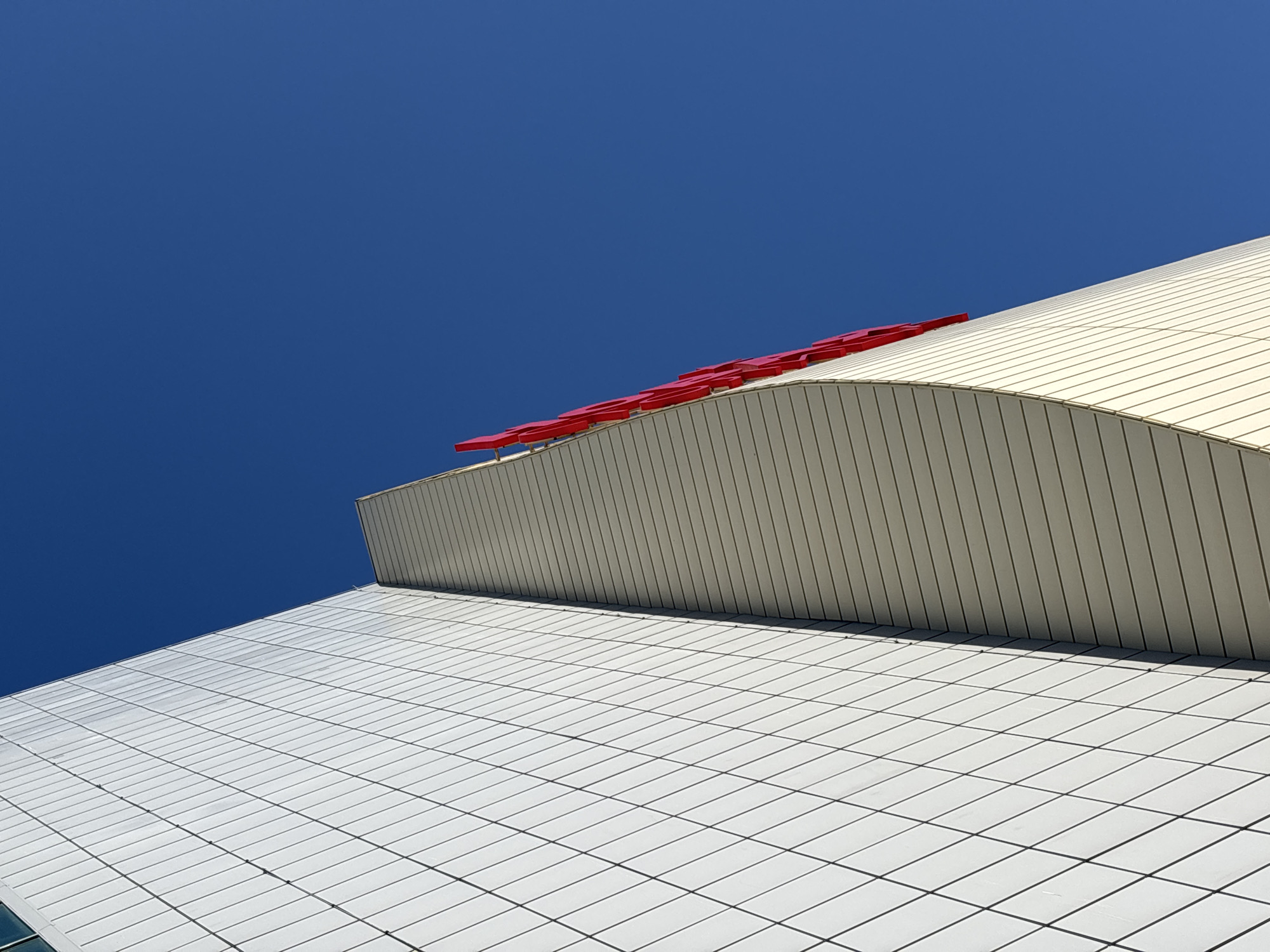 The image depicts an architectural view of the Perm Opera and Ballet Theatre in Perm, Russia, against a clear blue sky. The low-angle shot emphasizes the distinct architectural features of the building, showcasing the upper section of the facade. A curved, horizontally ribbed surface sits atop a gridded lower section. A series of red, rectangular forms are positioned along the crest of the curved section. The overall impression is one of modern architectural design.