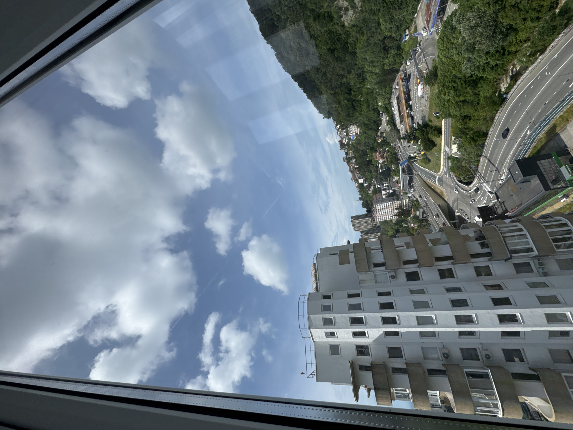 The image captures a view of a coastal city, likely Sochi, Russia, from an elevated perspective, possibly a high-rise building or a cable car. The foreground is dominated by the exterior of a white, multi-story building with distinctive curved balconies and numerous windows. To the right, a lush green mountainside rises steeply, dotted with houses and trees. A highway curves through the scene, with cars visible on its lanes. In the distance, other buildings and structures can be seen, contributing to the urban landscape. The sky is blue with scattered white clouds. The overall impression is of a sunny day in a city that combines urban development with natural mountainous terrain.