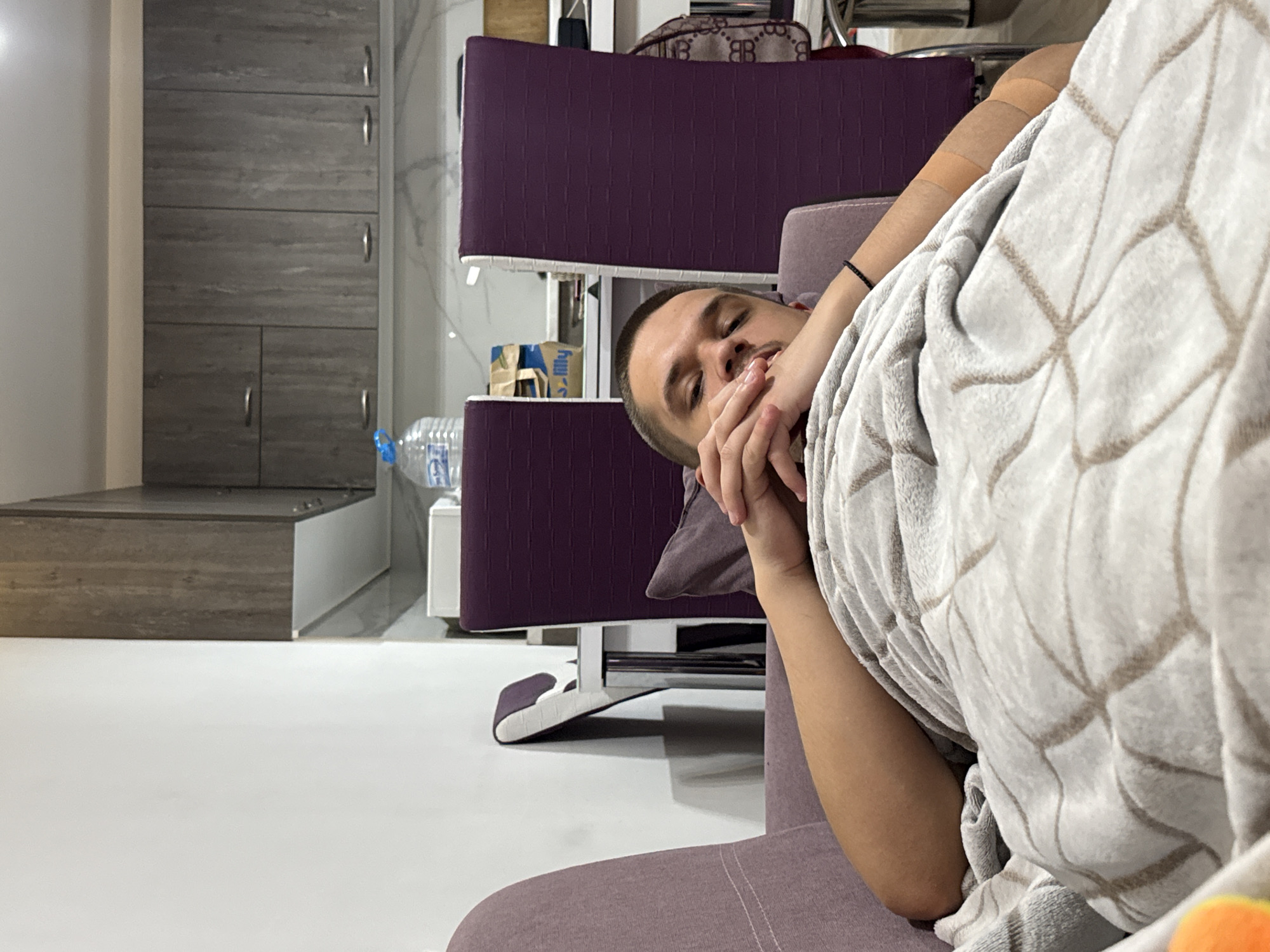 A man is seen from a tilted perspective, covered partially by a patterned, light-colored blanket while reclining in a purple upholstered chair. He is holding his hand near his mouth. Behind him, there's a multi-tiered purple structure, possibly shelving, and a water bottle resting on a surface. A gray cabinet with silver handles is visible on the left side of the frame, and the floor appears to be light-colored and reflective. The location is Plovdiv, Bulgaria.