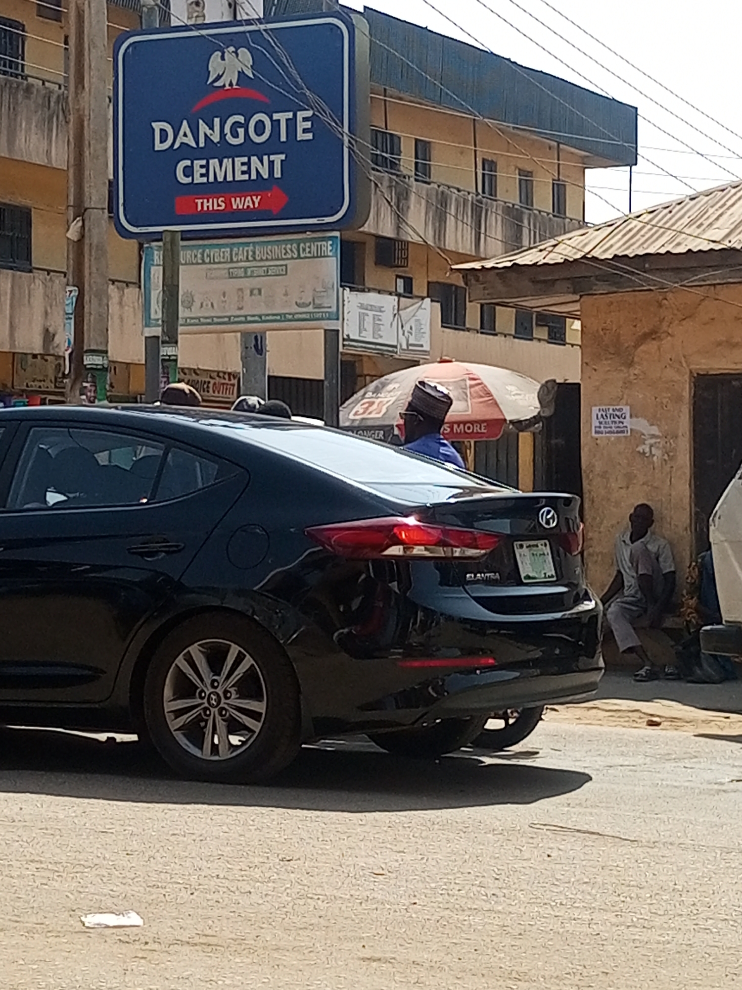The image depicts a street scene in Maiburiji, Nigeria. Prominently featured is a black Hyundai Elantra parked on the road. A directional sign for Dangote Cement is visible overhead. Two men wearing traditional hats are standing near the car. The background includes a building with several signs. A man sits on the ground near a building.