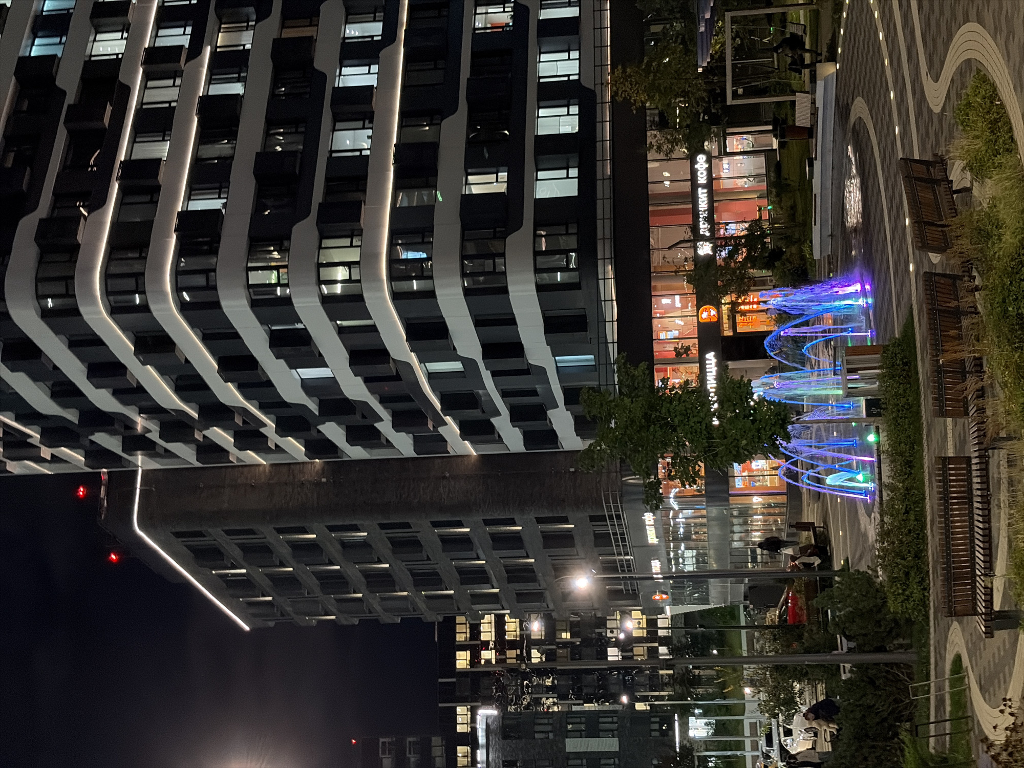 The image depicts a nighttime scene in Moscow, Russia, featuring a modern urban environment with several large residential buildings. In the foreground, a colorful illuminated fountain sprays water, creating a vibrant display. Several benches are placed around the fountain and in the surrounding paved area, which has a decorative, wavy pattern. Lush greenery and trees are scattered throughout the scene, some illuminated by ground lights.
A mid-rise building with a dark facade and white curved accents runs along the right side of the frame, its windows illuminated. To the left of this, a taller, darker building with vertical lighting lines and red lights on its roof is visible. Further in the background, more apartment buildings with illuminated windows can be seen, suggesting a densely populated residential area.
At the base of the buildings, a commercial area with storefronts is apparent. Signs with Cyrillic text, including "АРЕКИТ КОФЕ" (Areekit Coffee) and "ПИЦЦА" (Pizza), are visible on the storefronts. People are present in the scene, including some sitting on benches near the fountain and others walking in the background, one individual sitting on a swing set to the far right. Vehicles, including cars and what appears to be a red sports car, are parked or driving in the background. The overall atmosphere is one of a lively, contemporary urban setting at night.