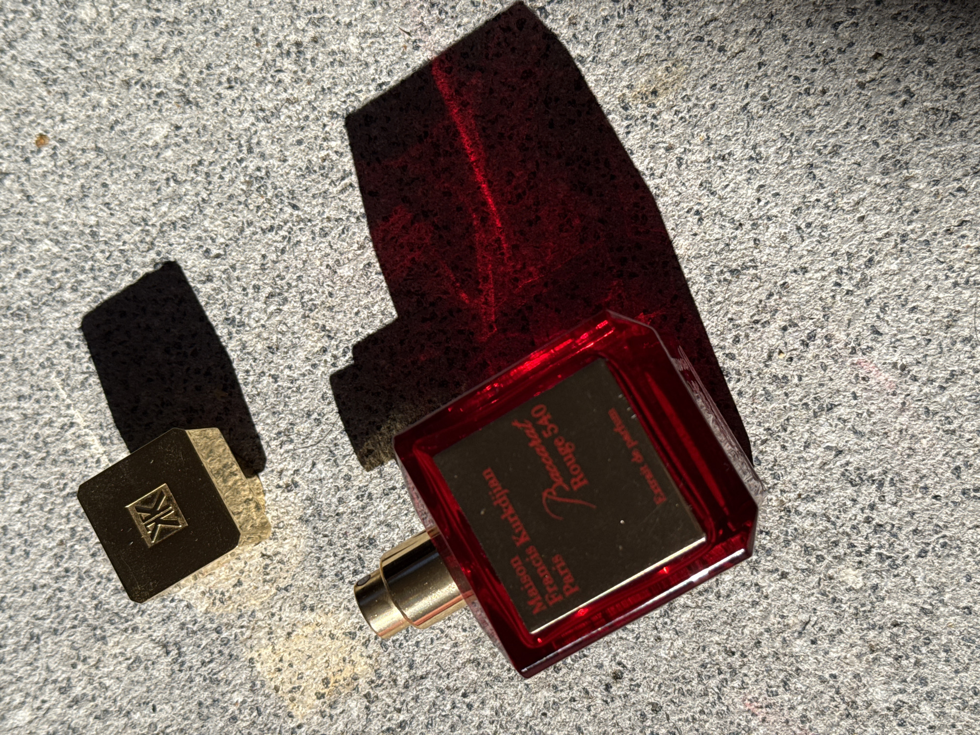In Bad Sachsa, Germany, on a sun-drenched, coarse-grained concrete surface, a luxurious red perfume bottle is positioned. The bottle, with its gold cap and metallic label, displays the text "Maison Francis Kurkdjian Paris" and "Baccarat Rouge 540 Extrait de parfum." A separate gold cap, embossed with the initials "KK" inside a square, rests nearby. The harsh sunlight casts distinct shadows of both the bottle and its cap onto the textured ground, creating areas of deep shade and highlighting the reflective qualities of the glass and metal. The scene is devoid of human presence.