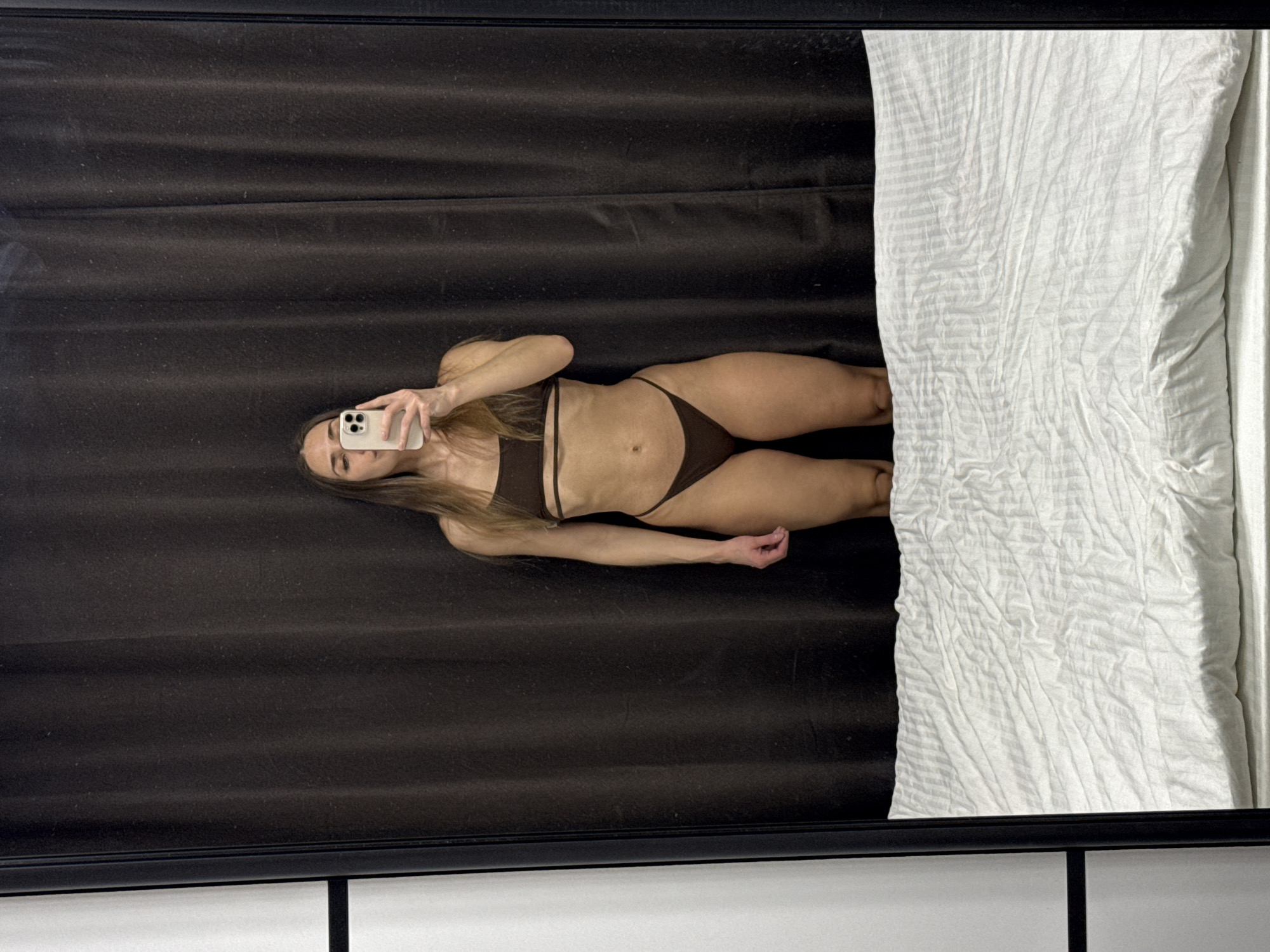 The image is a mirror selfie taken by a woman indoors, rotated 90 degrees counter-clockwise, making the woman appear horizontal.
**Subjects:** The primary subject is a woman with long, light brown hair, lying on her back as seen in the reflection. She is wearing a dark brown two-piece bikini or underwear set, consisting of a bandeau-style top and high-cut bottoms. Her left arm is extended upward, holding a white smartphone to capture the selfie. Her face is partially obscured by the phone, revealing her eye and forehead. Her right arm is resting by her side.
**Objects:** The woman is holding a white smartphone, likely an iPhone, with a visible camera array on the back. The reflection is captured in a large, dark-framed mirror. On the right side of the mirror's reflection, a portion of a bed with white, wrinkled sheets is visible. The background behind the woman in the reflection appears to be dark brown, possibly curtains or a wall, with subtle horizontal folds or texture. The bottom edge of the mirror frame is black.
**Activity or event taking place:** The woman is actively taking a selfie using her smartphone in front of a mirror.
**Notable details:** The image perspective is from inside the mirror, capturing the woman's full body. The white bed sheets provide a strong contrast to the dark brown background and the woman's attire. The woman has a lean physique. The image is rotated, giving an unusual orientation to the scene. There is no visible text in the image. The provided location context is Lyubertsy, Russia, but this is not discernible from the visual content of the image. The time of day and specific emotions are not clearly indicated.