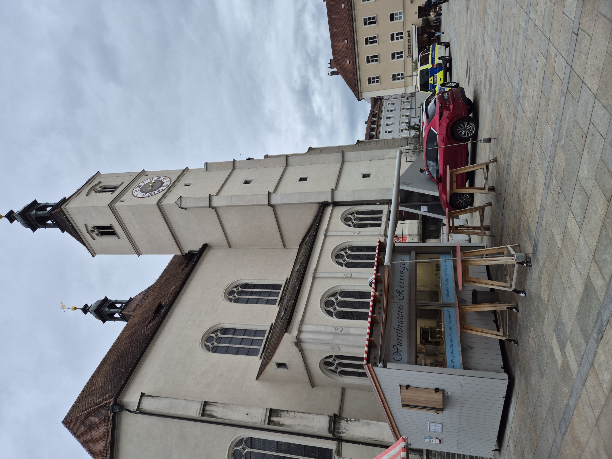 The image depicts a cloudy day in a European city square, likely Regensburg, Germany, given the architecture. Dominating the left side of the frame is a large, cream-colored church with a prominent bell tower featuring a clock face. The church has a steep, tiled roof and arched windows with Gothic tracery. In front of the church, a small, light-colored food stand with a striped awning is set up, advertising "Wurstbraterel Reisinger." Several wooden high-top tables with red tops are placed in front of the stand.
To the right of the food stand, a red SUV is parked. Behind the SUV, a yellow and blue police vehicle is visible, its lights flashing. Further in the background, other buildings with tiled roofs and multiple windows line the square. A sign on one of these buildings reads "COFFEE FELLOWS." A few people are scattered throughout the scene, including one person near the red SUV and two near the police vehicle, suggesting a public space. The square is paved with large, square stone tiles, and a dark grey stripe runs across the foreground. The overall atmosphere is subdued due to the overcast sky.