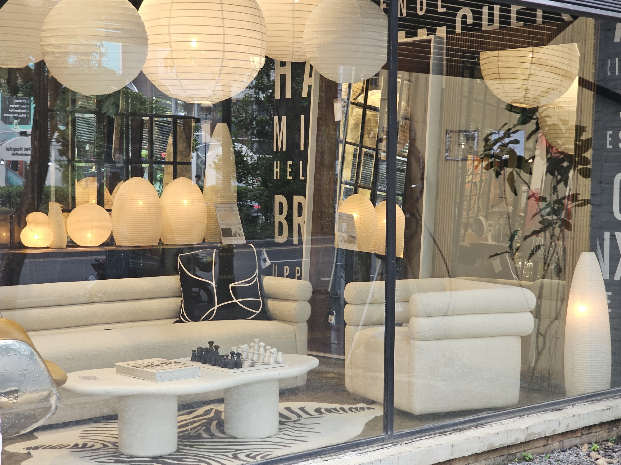 A street-level storefront in Bangkok, Thailand, displays a modern interior with a beige modular sofa and armchair arrangement, a white oval coffee table with a book and a chessboard set up, and various paper lantern-style light fixtures. The storefront has a large glass window, reflecting the outdoor environment including trees and buildings. The interior features a striped rug underneath the furniture. The lighting inside is warm and diffused, emanating from the paper lanterns. Behind the furniture, the background is somewhat blurred, showing more interior elements and reflections of the street. The overall impression is a clean and contemporary retail space.