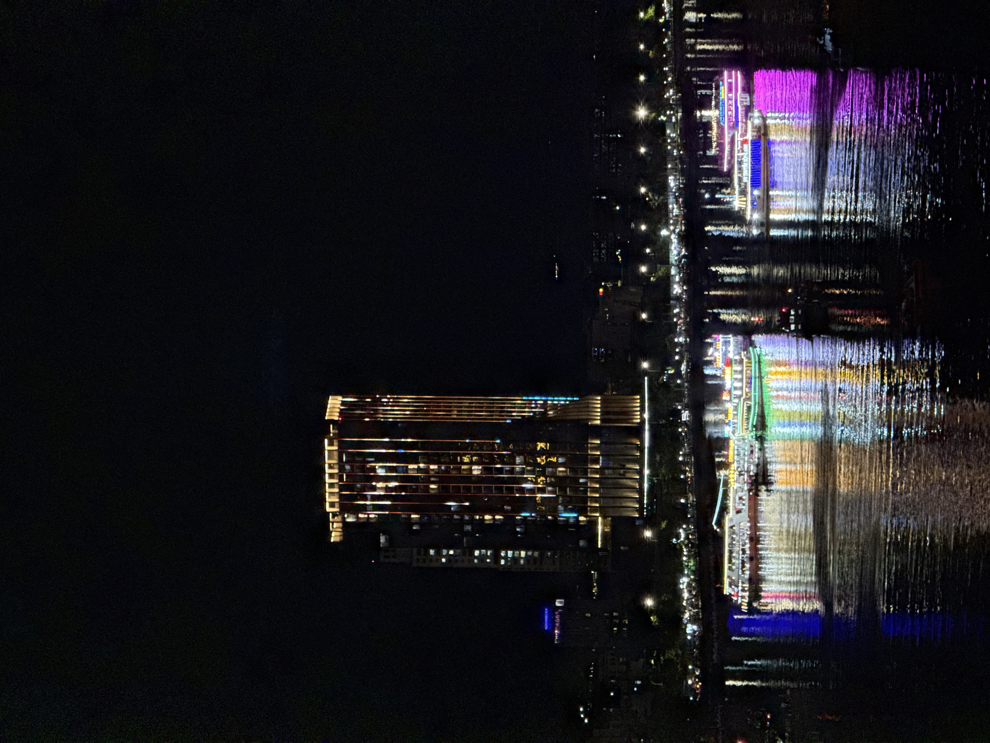 The image depicts a nighttime view of the Han River in Đà Nẵng, Vietnam. In the background, a tall, modern skyscraper stands illuminated with vertical lines of warm yellow and orange lights, reflecting on its glass facade. To the left of this main building, another, less illuminated skyscraper is visible, with some windows showing light. Along the riverbank, a string of lights indicates a promenade or road lined with trees and streetlights.
The foreground is dominated by the dark water of the Han River, which is aglow with the colorful reflections of multiple tour boats. These boats are brightly lit with various colors, including green, white, blue, and pink. Several boats are actively moving on the river, their lights creating shimmering trails on the water's surface. The overall scene conveys a vibrant and lively urban nightscape.