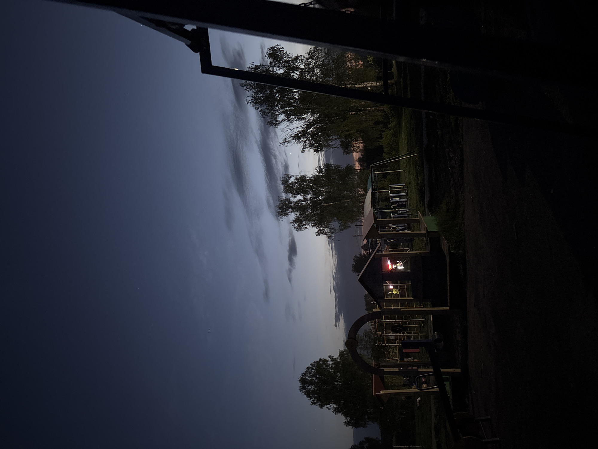Captured at night in Фоки, Russia, the image presents a playground scene. In the foreground is the playground with partially visible climbing equipment and structures, obscured by the darkness. A building, possibly a small house or clubhouse, is situated within the playground, illuminated with interior lights. Beyond the playground, trees are silhouetted against the sky, which transitions from a darker blue to lighter hues with visible clouds. The overall light level is low.