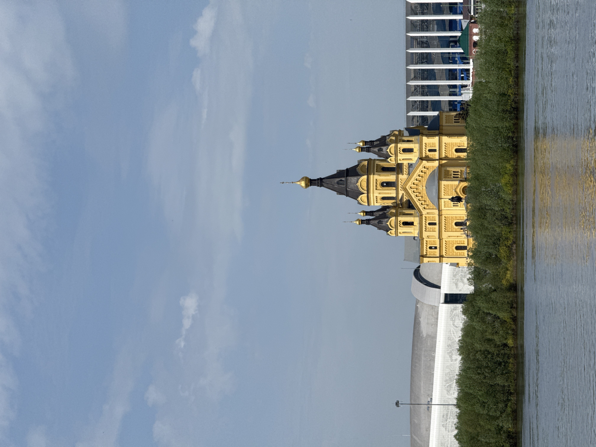 The image depicts the Alexander Nevsky Cathedral in Nizhny Novgorod, Russia, situated along a body of water, likely the Oka or Volga River. The cathedral, characterized by its golden domes and yellow facade, stands next to lush greenery along the water's edge. A modern, white architectural structure, possibly a stadium or concert hall, is partially visible in the foreground. In the background, parts of modern glass and steel buildings can be observed. The sky is light blue with scattered clouds. The overall scene appears to be a calm, sunny day.