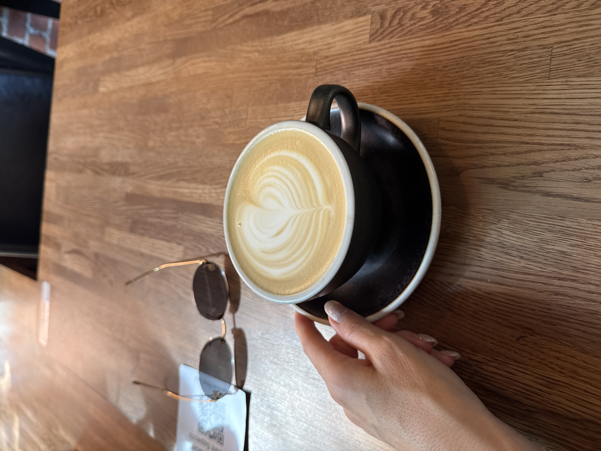 A close-up, high-angle shot shows a woman's hand with glittery nail polish reaching for a latte with latte art in the shape of a heart, placed on a saucer. The coffee cup is black with a white rim. In the background, a pair of sunglasses rests on a wooden table next to a piece of paper with a QR code. The setting is likely a cafe or restaurant. There is no indication that the scene is located in Moscow, Russia.