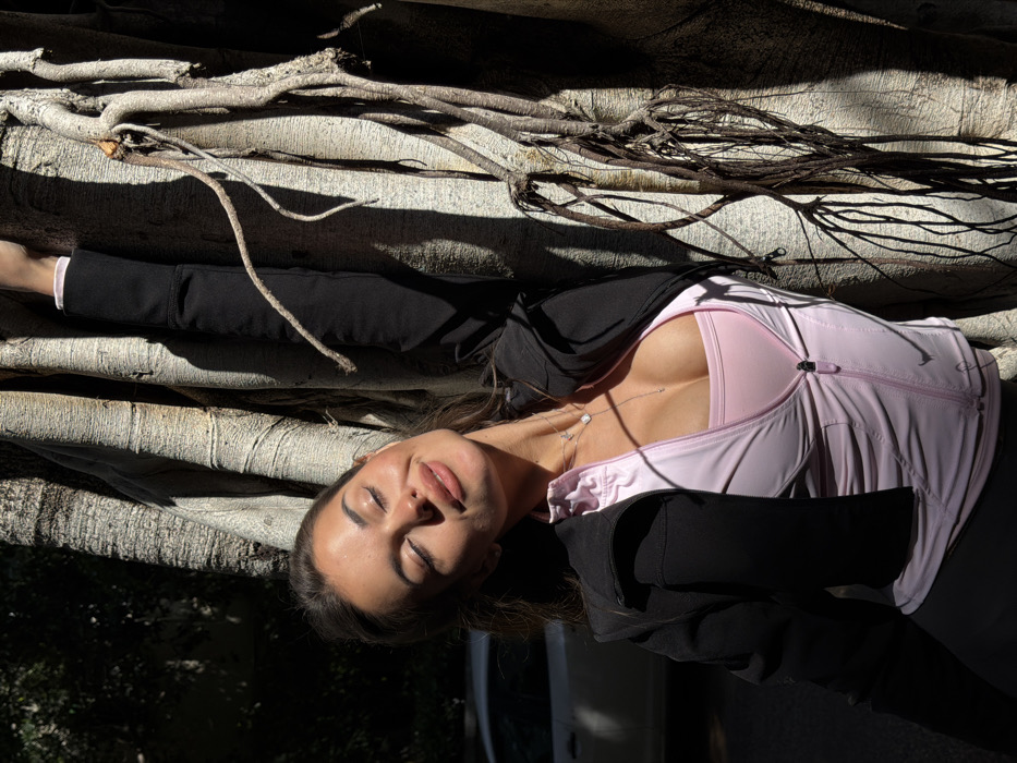 A single adult female is depicted in an outdoor, sunlit close-up against a large, multi-trunked Ficus tree. The subject, positioned on the left, has brown hair and light-to-medium skin, with her eyes closed and head tilted upwards, a slight smile on her lips. She is wearing a light pink sports bra beneath a partially unzipped light pink athletic jacket, layered with an open black cropped jacket. A silver necklace with a small pendant adorns her neck. Her right arm extends upward, interacting with the tree trunk. The tree's textured bark and numerous aerial roots dominate the right portion of the frame, casting distinct shadows from direct sunlight. In the blurred background to the left, a light-colored vehicle is partially visible. The scene is consistent with an urban outdoor environment in Tel-Aviv, Israel, where Ficus trees with prominent root systems are a common feature.