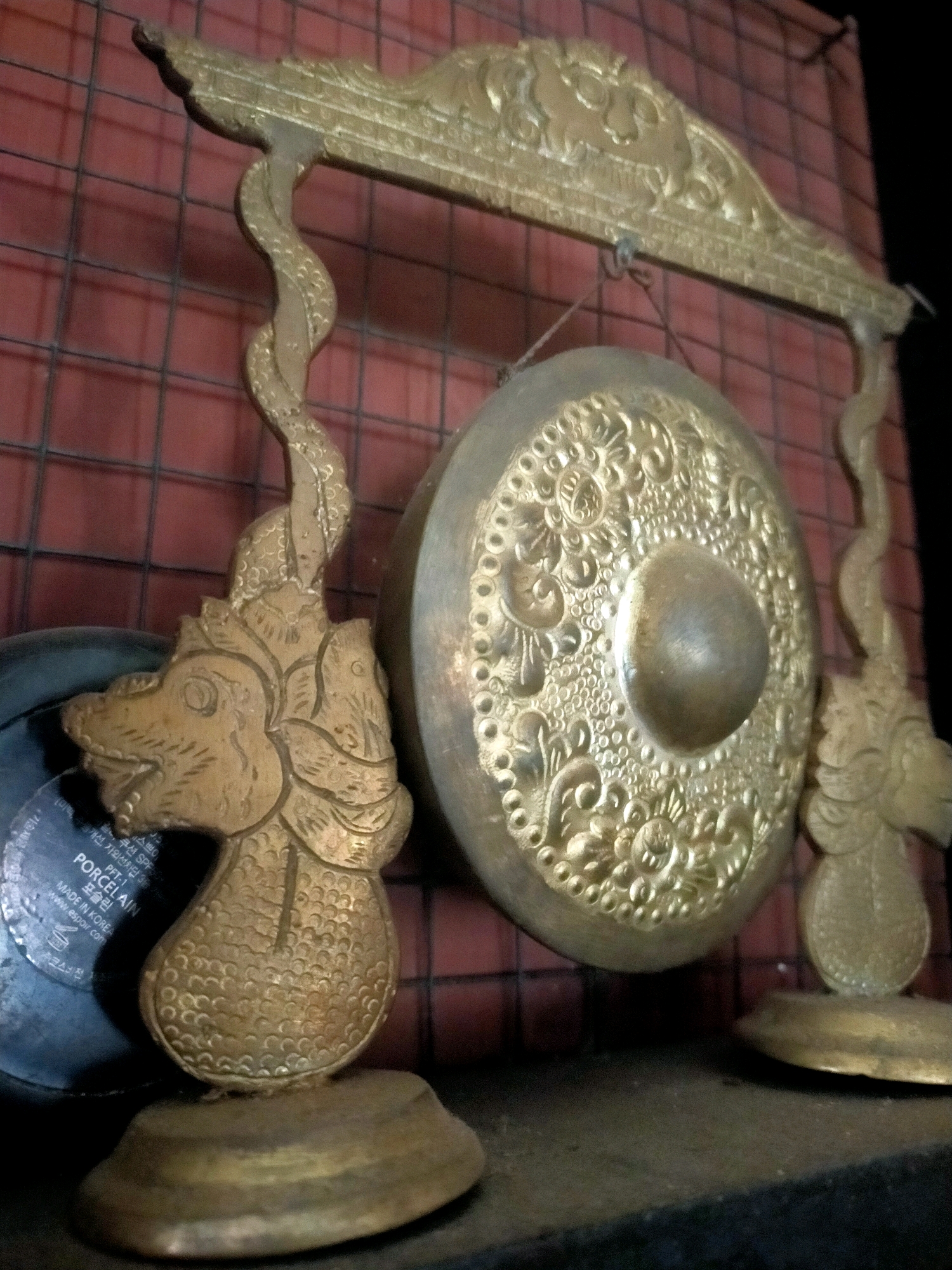 The image shows a close-up view of a decorative gong stand, likely made of brass or a similar metal, featuring ornate dragon head carvings supporting a decorative arched top. A circular gong with intricate embossed patterns hangs from the stand. The background appears to be a reddish-brown grid-like surface, possibly a brick wall with a wire mesh in front of it. To the left of the gong stand, partially visible, is a dark, circular object with some text printed on it, including the words "PORCELAIN" and "MADE IN KOREA". The scene is well-lit, highlighting the metallic sheen and detailed craftsmanship of the gong stand and gong. The overall impression is one of traditional craftsmanship and decorative artistry. The location in Karangsono, Indonesia, suggests this might be an artifact displayed in a home, cultural center, or shop specializing in Indonesian crafts.