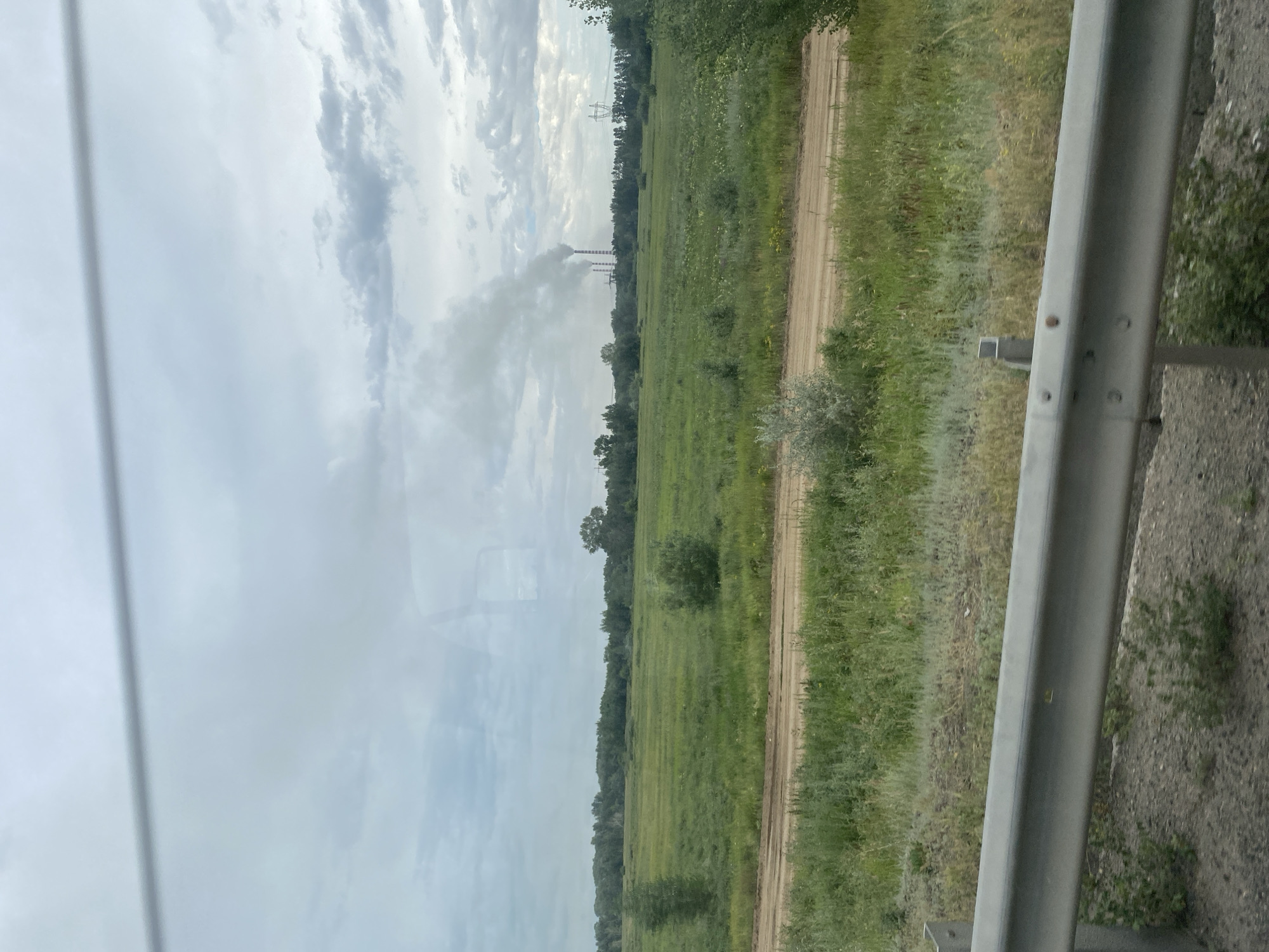 The image depicts a pastoral landscape with a power plant in the distance emitting smoke. The foreground features a dirt road and a guardrail, suggesting a roadside view. The sky is overcast with clouds, and the surrounding area is covered in green grass and scattered trees, indicating a rural or semi-rural setting within the Aqsu city administration of Kazakhstan. There are no people or specific landmarks visible that would allow for a more precise location within the city administration. The power plant with its smoking chimneys is the most prominent man-made structure in the scene.