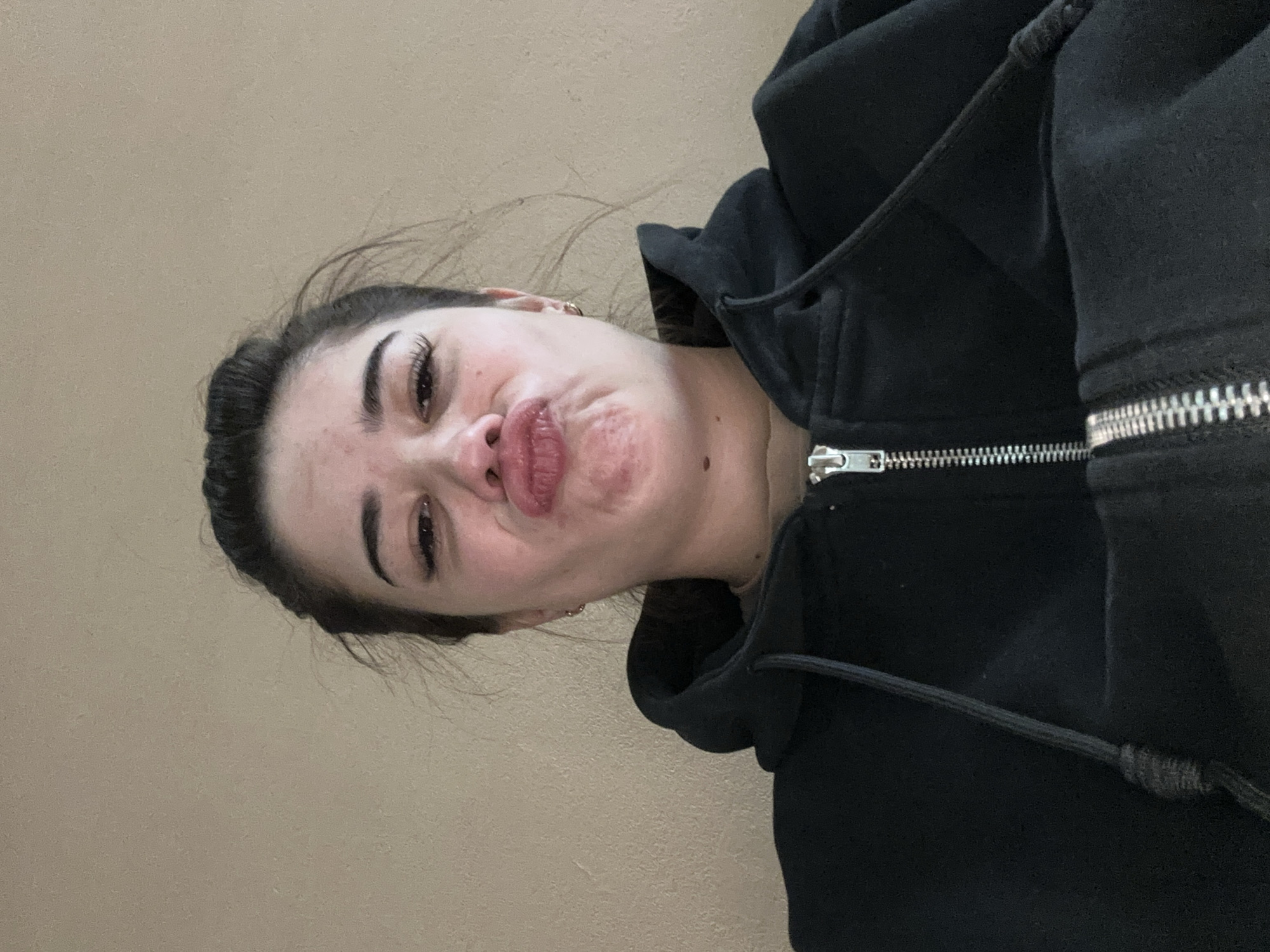 A young woman with dark hair pulled back in a bun and wearing a black zip-up hoodie is looking up at the camera with her lips puckered. She has long eyelashes and is wearing small hoop earrings. The background is a plain, textured wall. The image appears to be a selfie taken indoors. There is no indication of the city of Gavrilov Posad, Russia, in the image.