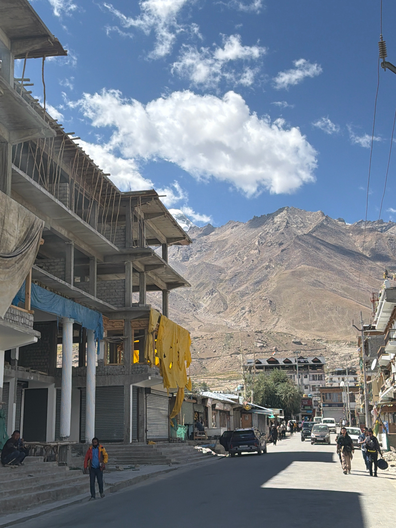 The image depicts a street scene in Padum, India, with a backdrop of rugged mountains. In the foreground, a building under construction is prominent on the left, with exposed concrete floors and supporting wooden structures. Tarpaulins, one yellow and one blue, are draped over parts of the building. A paved street runs through the center, with several vehicles including cars and a motorcycle parked or in motion. On the right side of the street, more buildings line the road, some appearing to be shops with closed shutters. Several individuals are visible walking on the street and sidewalks. The sky is blue with scattered white clouds, and the mountains in the background are rocky and arid. The overall scene suggests a developing area within a mountainous region, likely a town or village.