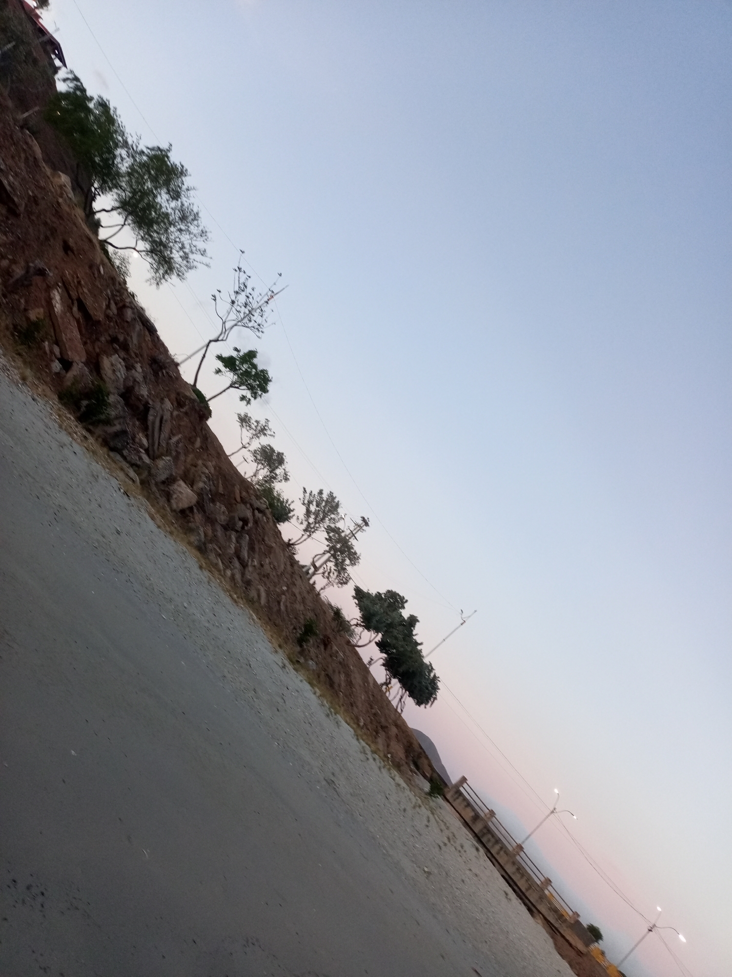 The image shows a road adjacent to a rocky slope in Juan Griego, Venezuela. Trees grow along the top of the slope. A bridge with railings and street lights is visible on the right side of the road. The sky is a gradient of light blue and pink, indicating either sunrise or sunset. The road surface appears to be asphalt or a similar material.