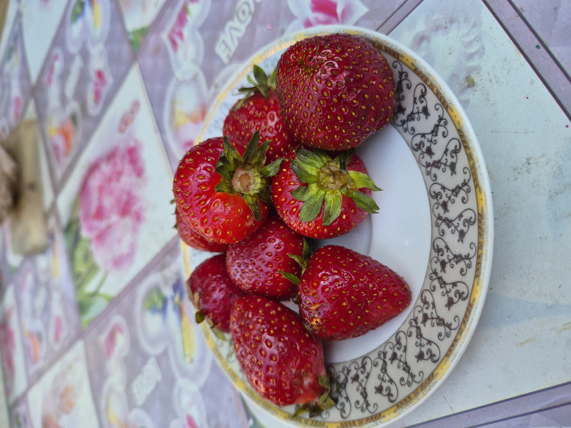 A close-up, vertical, eye-level shot shows a white plate with a gold and brown decorative rim filled with six bright red strawberries. The strawberries are plump and have visible seeds. The plate rests on a surface covered with a light-colored, patterned tablecloth. The pattern features squares with faded floral designs and the word "LOVE" repeated. In the blurred background, another wooden surface is visible. The lighting suggests an outdoor setting.