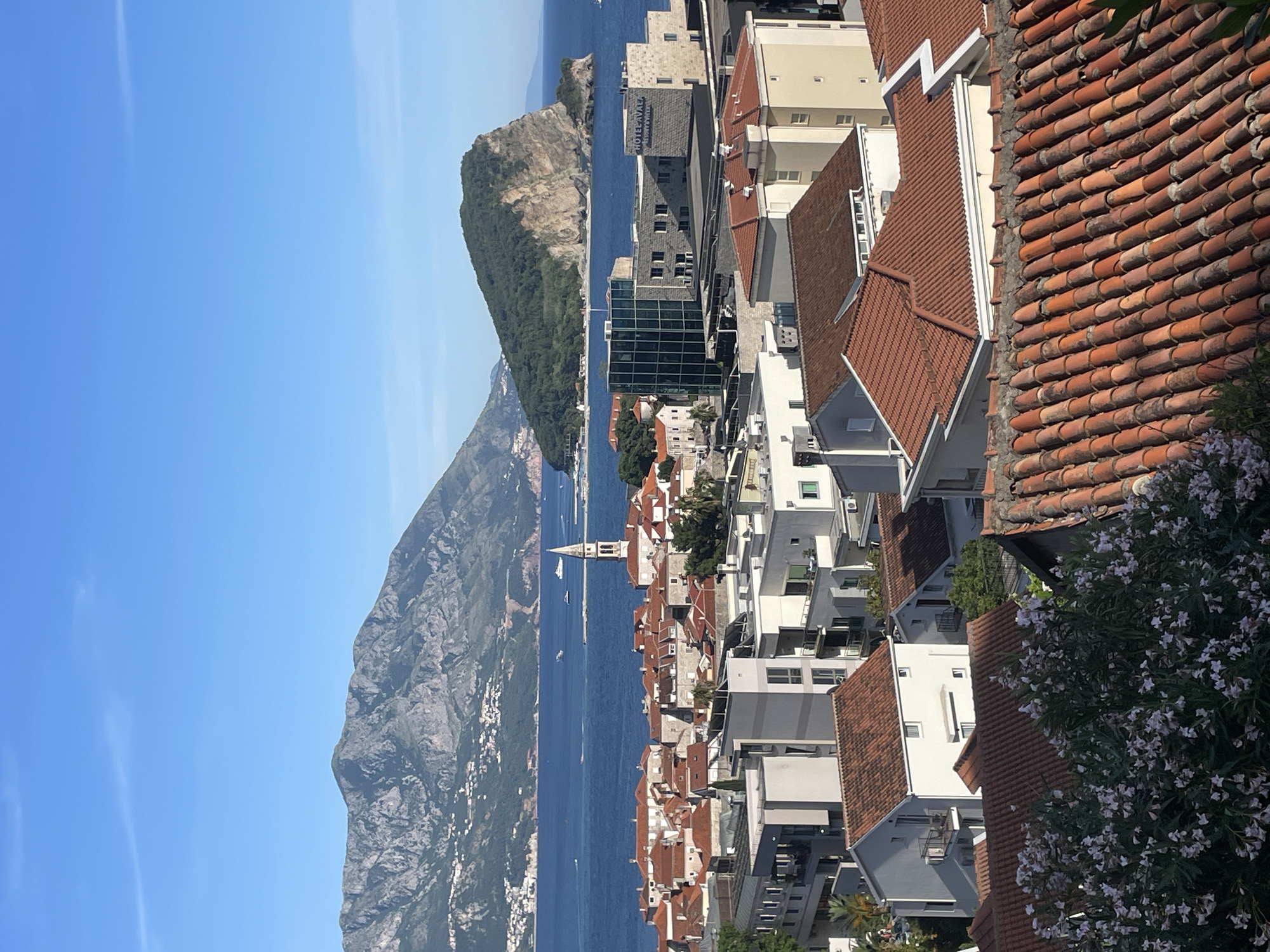 The image shows a panoramic view of Budva, Montenegro, a coastal city. In the foreground, the red-tiled roofs of buildings are visible, indicative of the area's traditional architecture. Modern buildings are interspersed among the traditional structures, including the Hotel Avala Resort and Villas. In the middle ground, the Adriatic Sea stretches out, separating the mainland from Sveti Stefan, a small islet connected to the coast by a narrow isthmus. The sea is dotted with small boats. Behind Sveti Stefan, a steep, mountainous landscape dominates the horizon. The sky is clear and blue.