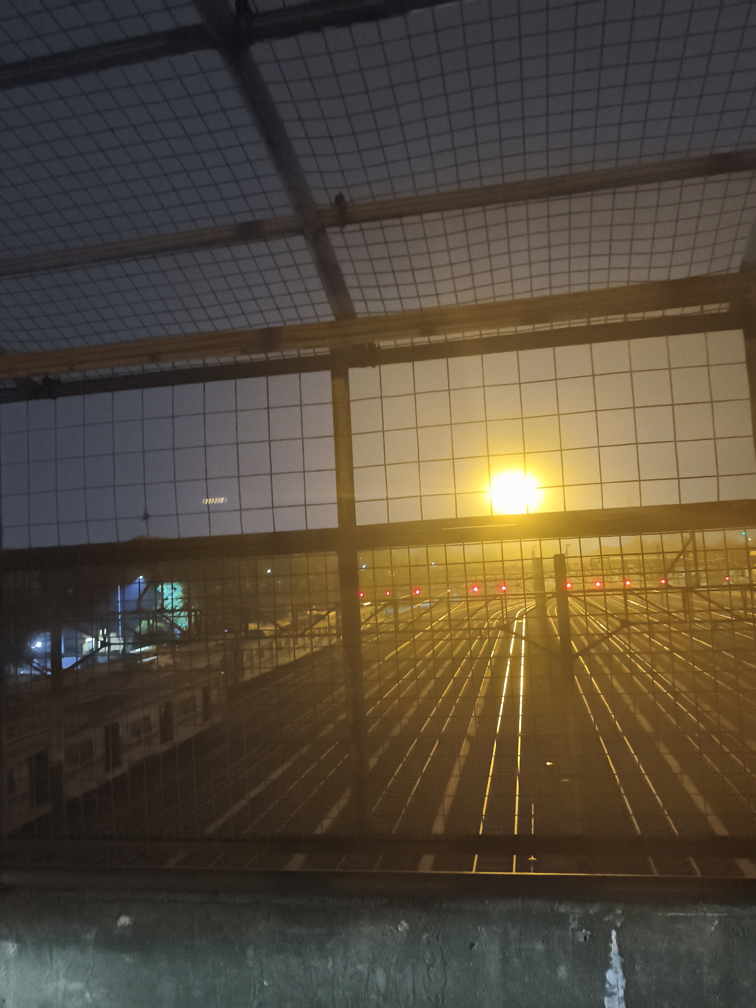 The image depicts a railway yard at dusk or dawn, possibly at Depok Station in Depok, Indonesia. The foreground is dominated by a wire mesh fence and a metal railing, suggesting the photo was taken from an elevated vantage point, such as a pedestrian overpass. Beyond the fence, numerous train tracks stretch into the distance, illuminated by the warm glow of what appears to be artificial lighting. A bright, diffused light source, likely a streetlamp or floodlight, is visible in the sky, casting long, diagonal shadows across the tracks. Several red signal lights are also visible along the tracks, indicating railway operations. In the mid-ground, the side of a train is partially visible, suggesting it is parked at a platform or in a maintenance area. The sky is hazy, contributing to the soft, atmospheric lighting. The overall impression is of a functional and active railway infrastructure scene during a transitional time of day.