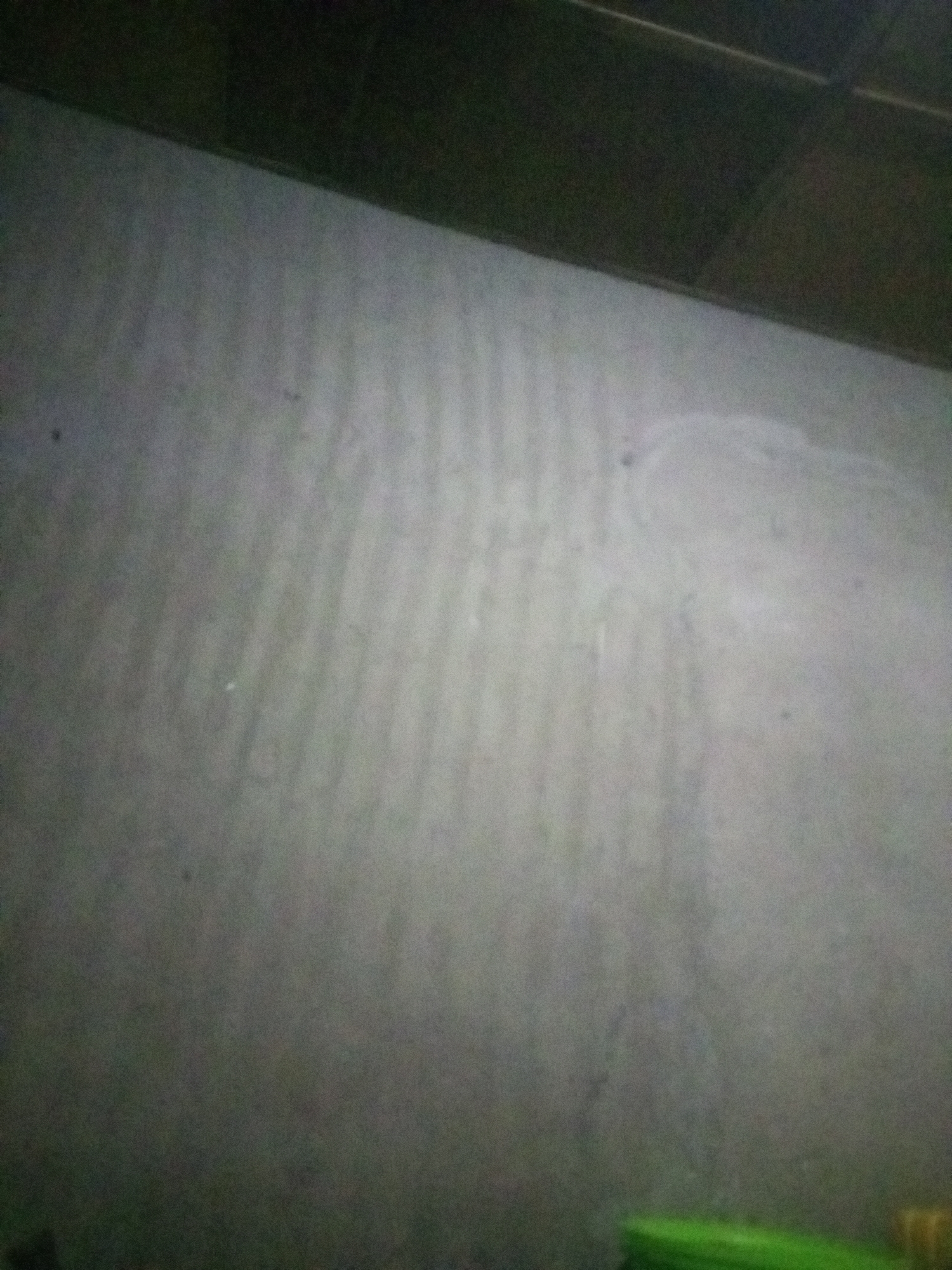 The image displays a dimly lit, textured wall with faint vertical lines or ripples, possibly indicating a concrete or plaster surface. The top portion of the image is dark, suggesting a ceiling or overhead obstruction. The overall impression is of an interior space, but no specific details about the city or location are discernible. There are no people or identifiable objects present in the frame.