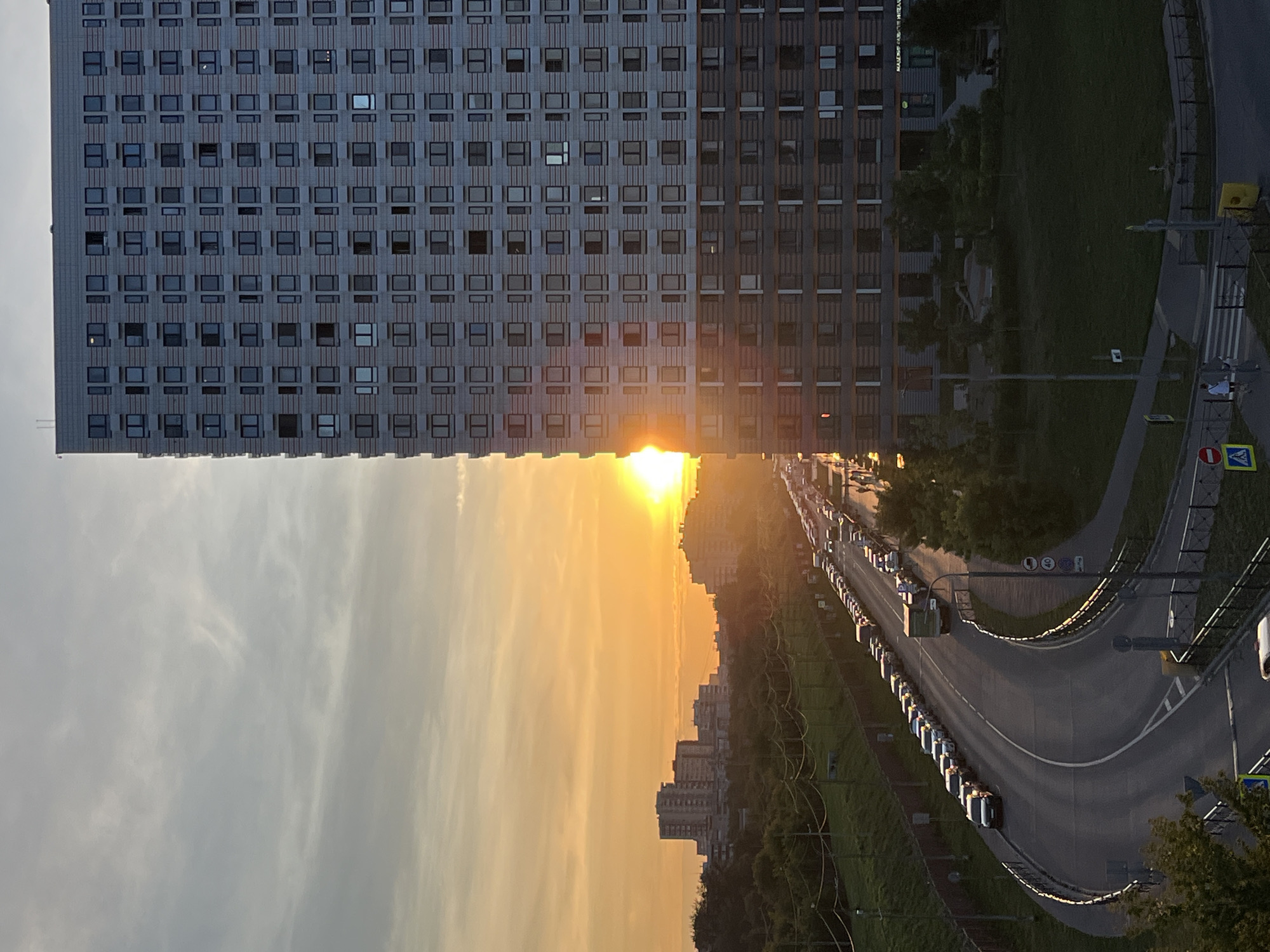This is a photograph taken from a high vantage point, looking east towards a sunset in Moscow, Russia. On the right side of the image is a tall, modern residential building with a grid of windows. The setting sun is partially obscured by this building, casting a warm orange glow across the sky. In the distance, a cityscape of other buildings and trees is visible. A curved road runs in front of the residential building, with a long line of cars parked along its side, suggesting a busy urban environment. The road is bordered by a grassy embankment and a fence. Further down the road, a truck is visible, and a few people are walking on a sidewalk. The overall impression is of a typical urban evening scene with the dramatic backdrop of a sunset.