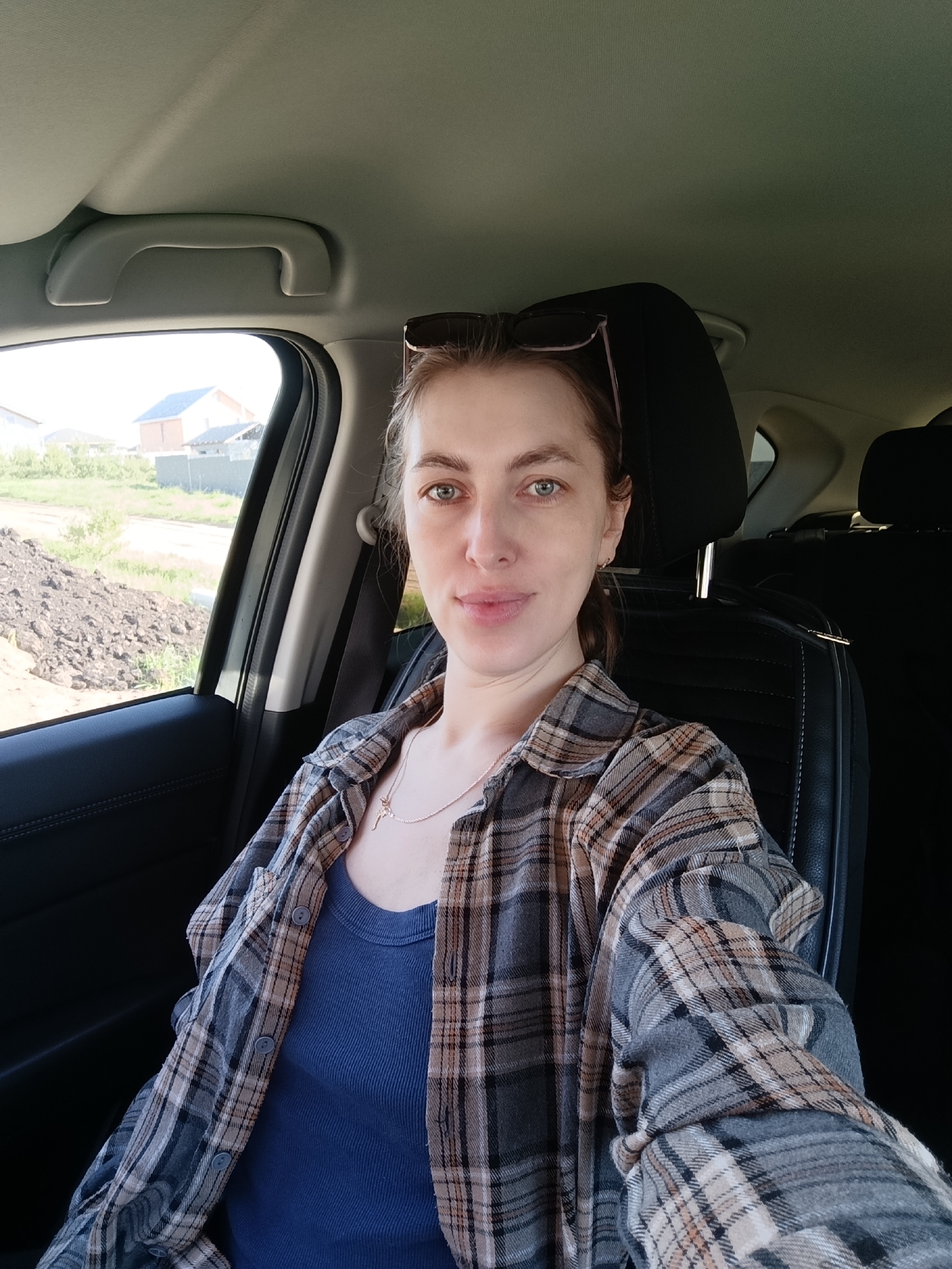 A woman with light skin, light brown hair with sunglasses perched atop her head, and green eyes is sitting in the driver's seat of a car. She's wearing a blue tank top and a plaid shirt. A silver necklace is visible around her neck. The car seat is black leather.
Outside the car window, there's a view of a landscape with a pile of dirt or construction material, some greenery, and a house in the background. The house is a light color with a blue roof.
Based on the location provided, this scene is situated in Chaikovsky, Russia.