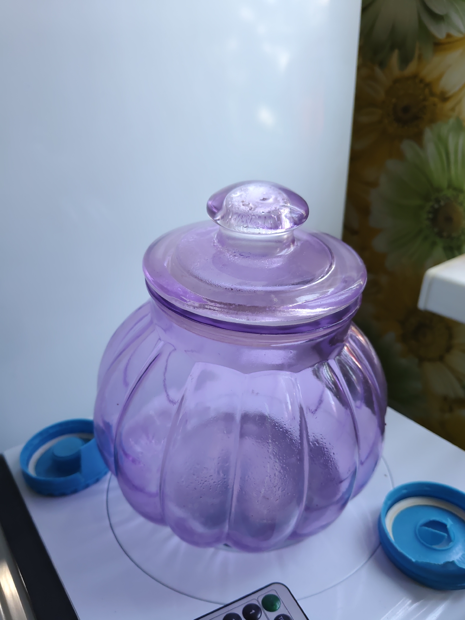 A purple glass jar with a lid sits on a white surface. To the left and right of the jar are two blue caps, likely from other containers. In the background, a white object is on the left and a patterned curtain with flowers is on the right. The scene is indoors and appears to be a home environment. There are no people present. The lighting is natural, coming from a window or overhead source. The image was taken in Rostov-on-Don, Russia.