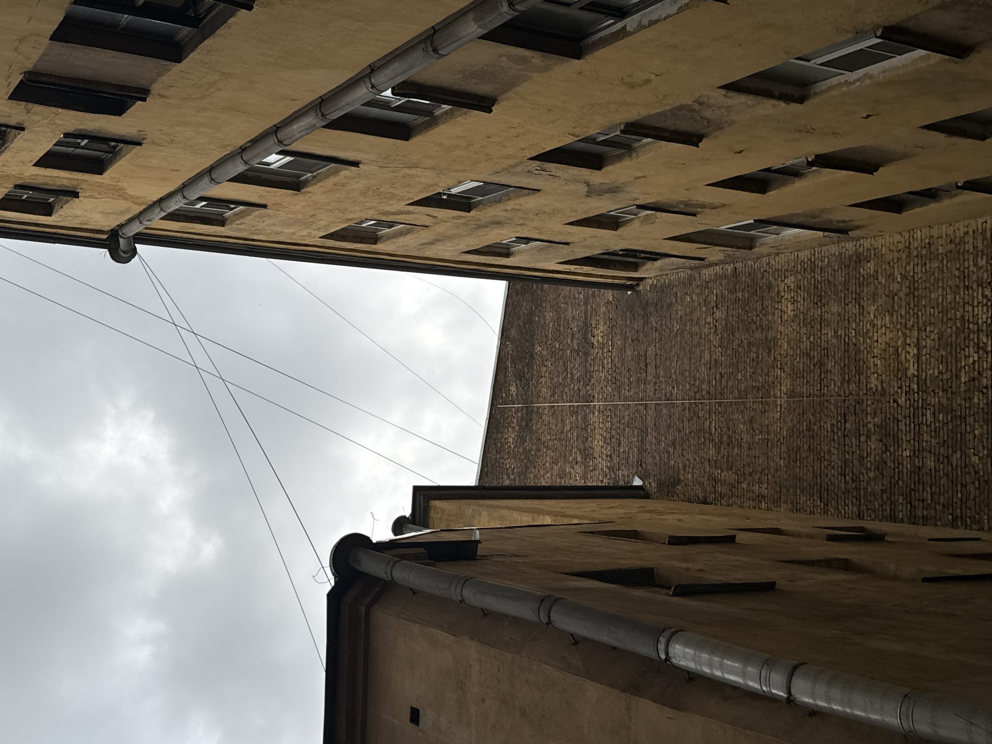 The image is an upward-facing shot of a narrow courtyard in Saint Petersburg, Russia, between two tall, old buildings. The building on the left is a tan stucco structure with a dark roofline and a large, silver downspout running vertically along its side. The building on the right is a yellow stucco structure with a series of evenly spaced windows and a similar silver downspout. The background is a brick wall. The sky is overcast and grey. There are power lines crisscrossing the sky. No people or animals are visible. The scene appears to be a residential area.