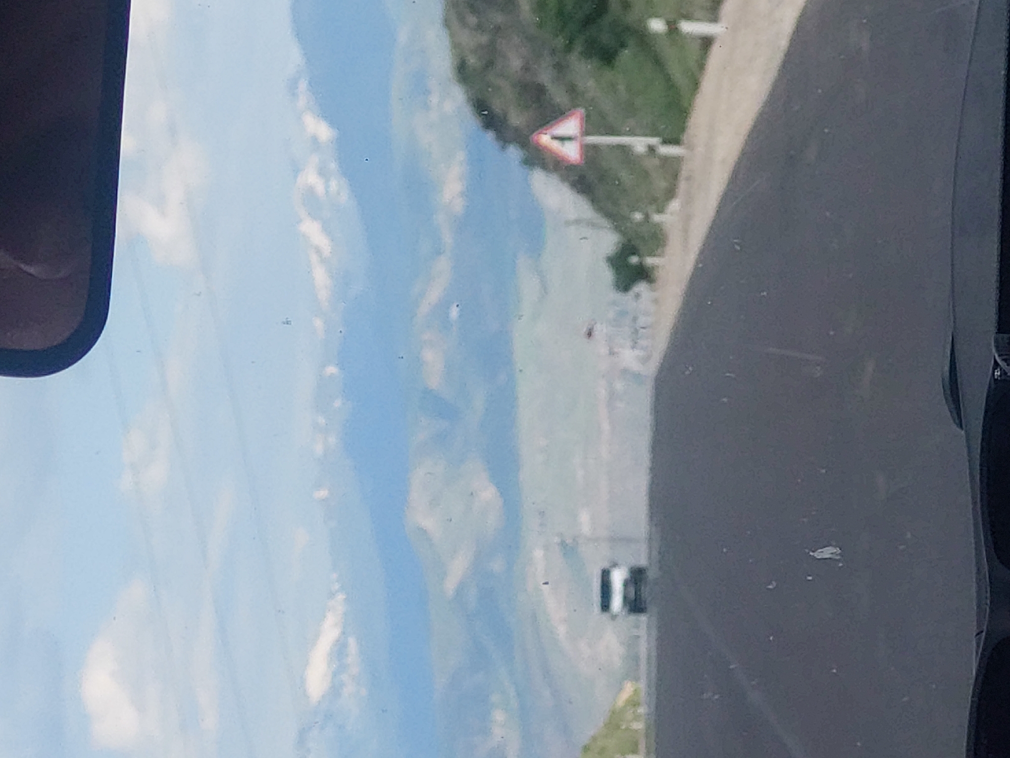 The image, oriented correctly, depicts a scenic view from inside a vehicle looking out over a mountainous landscape. On the far left, a portion of the vehicle's side mirror is visible. The right side of the frame shows a dark interior part of the vehicle, possibly a dashboard or window frame. The car window itself is speckled with small dust particles or water spots.
A bright blue sky with scattered white clouds dominates the upper half of the image. Below, a vast valley unfolds, flanked by multiple layers of green mountains and hills stretching into the distance. A dirt or gravel road runs along the right side of the frame, winding through the natural terrain.
In the mid-ground, a red-bordered white triangular warning sign with a black horizontal bar is prominently displayed on a white pole, positioned on what appears to be an embankment overlooking the valley. Further into the distance, within the valley, faint outlines of structures, possibly a remote settlement, can be seen. A small, light-colored vehicle, likely a car or van, is also visible on the road in the distant part of the valley.
The scene captures a daytime journey through a rural, mountainous region, consistent with the location context of Yelpin, Armenia. The weather appears clear and sunny. No explicit text is visible on the sign or elsewhere.