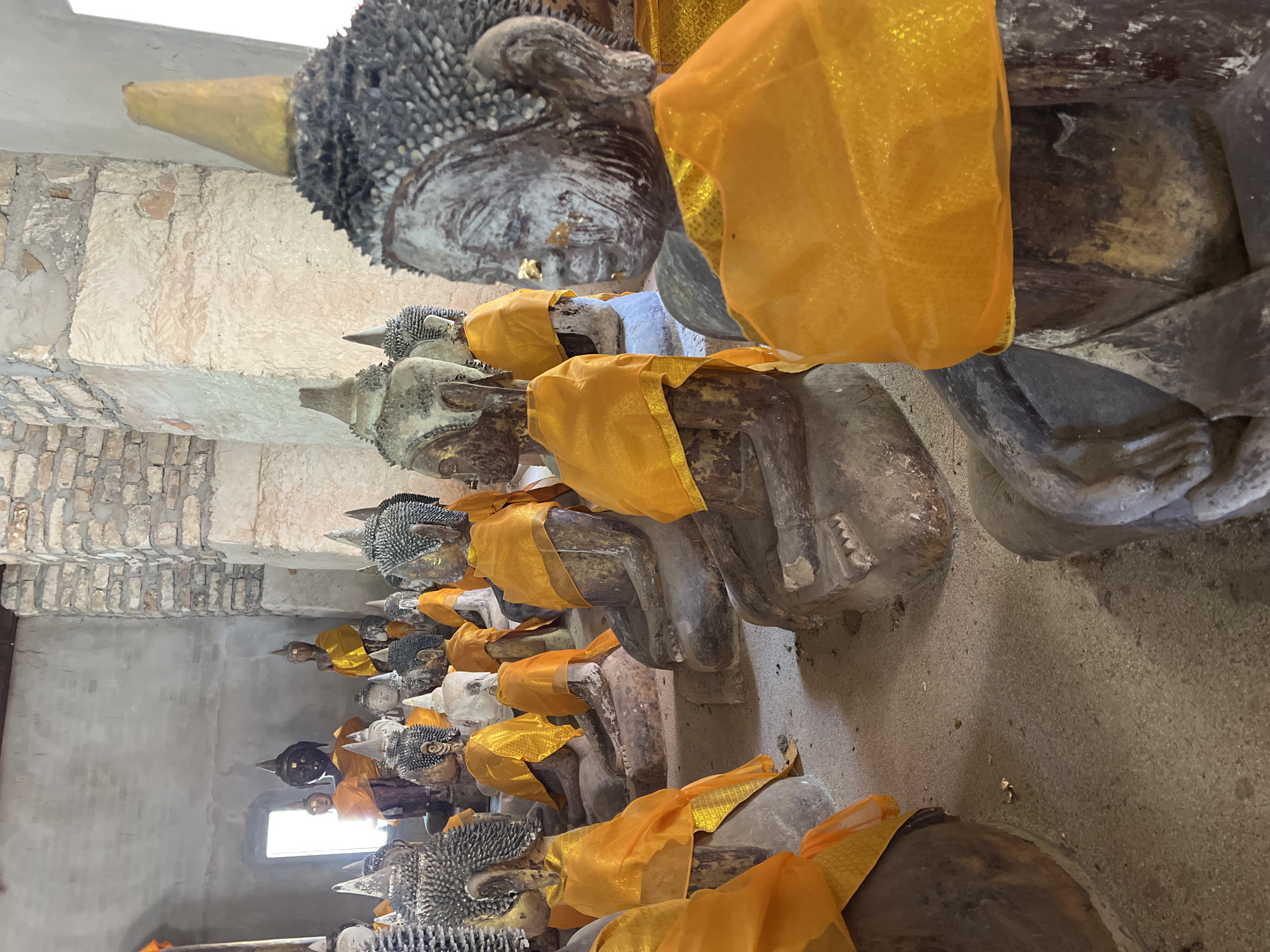 The image depicts a row of reclining Buddha statues inside a building in Baan Harn, Thailand. The statues are made of stone and are draped with orange fabric. The building's walls are made of brick. A window is visible at the far end of the room. The statues are arranged closely together along the length of the room. The light comes from the window at the end of the row.