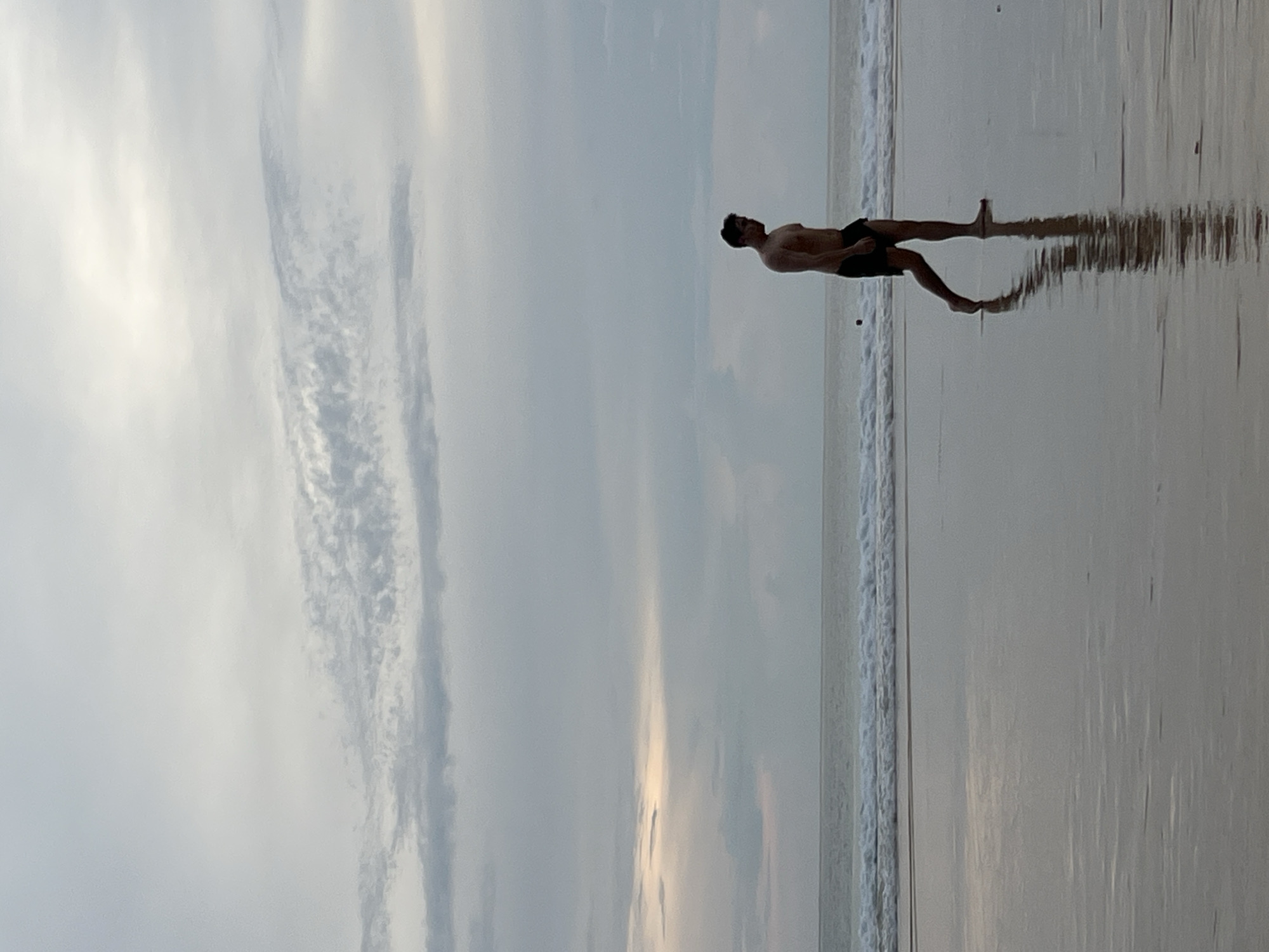 A shirtless man in black swim trunks walks along a wet sandy beach during overcast conditions. The man, in profile, faces left, his reflection visible on the wet sand. In the background, the ocean meets the sky with gentle waves. The sky is filled with scattered clouds, with a hint of light breaking through on the left horizon. The scene appears to be at Bang Tao Beach in Choeng Thale, Thailand.