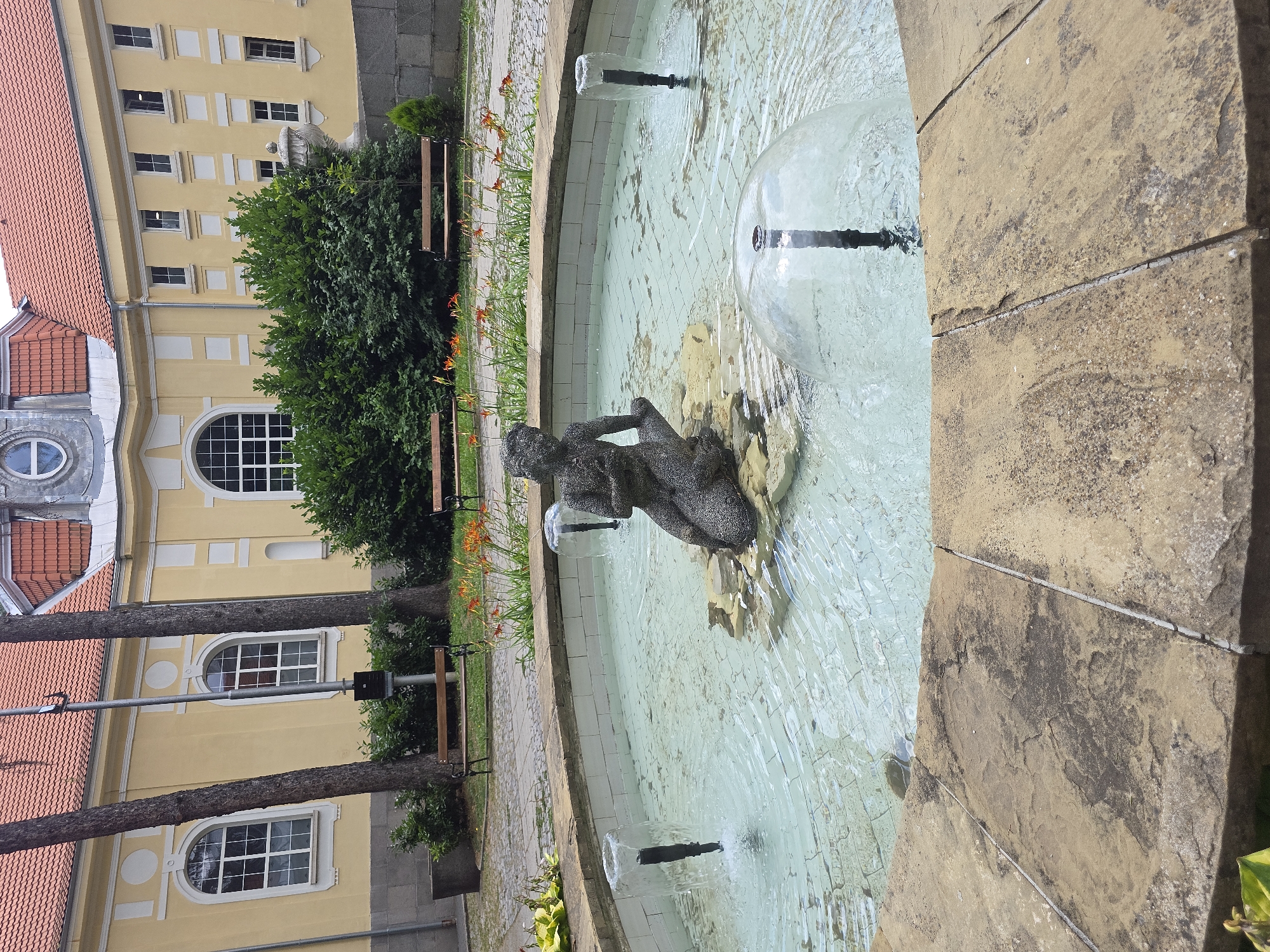 A circular fountain with a stone border is in the foreground. The fountain contains a stone sculpture of a crouching figure, possibly a mermaid, and three water jets, one of which is creating a dome of water. In the background, a yellow building with a red-tiled roof and arched windows is visible, along with trees, bushes, and two wooden benches.