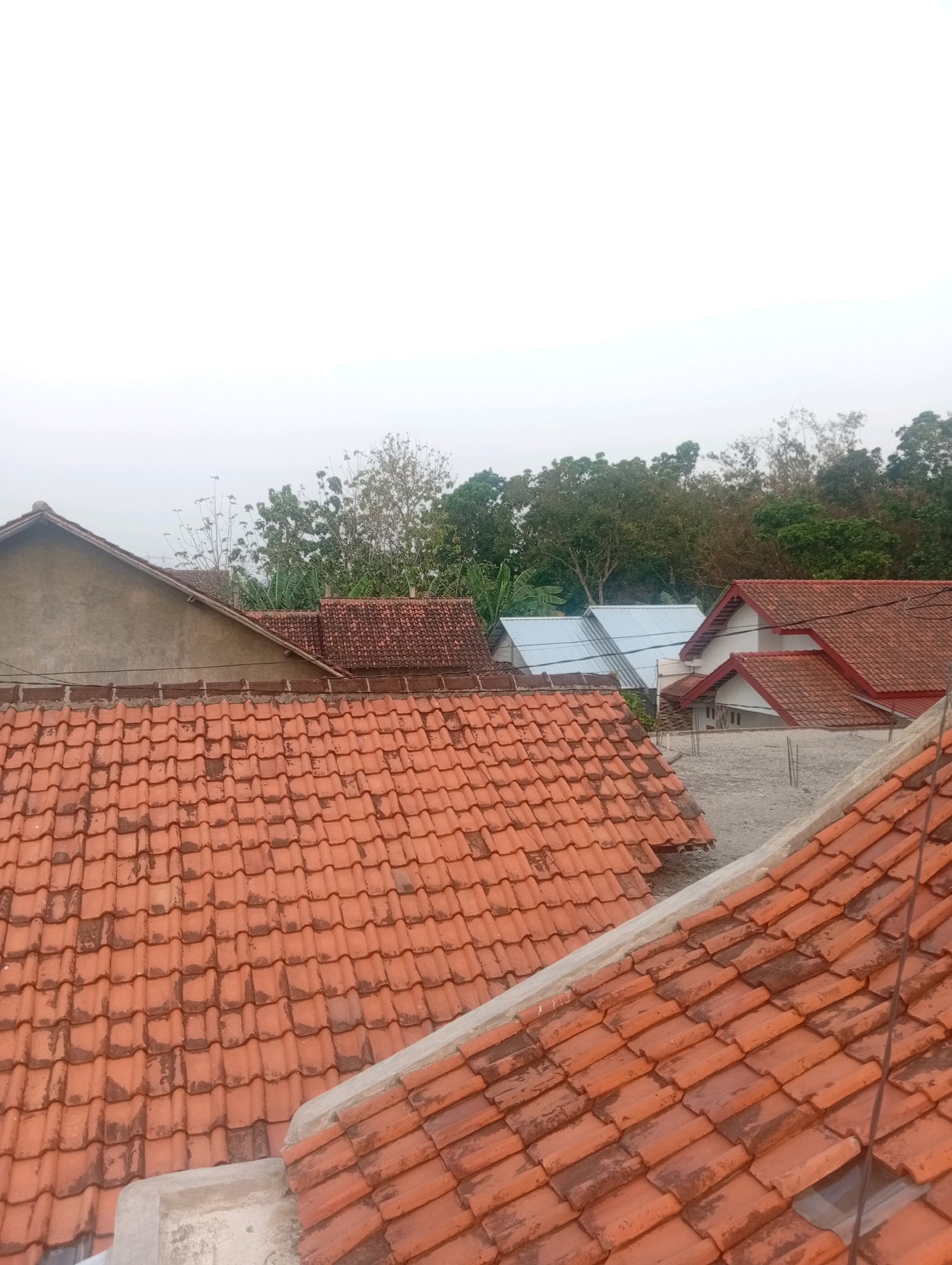 The image depicts a cluster of houses with red tiled roofs, situated in the Kebandingan area of Indonesia. The foreground is dominated by the textured surface of terracotta roof tiles. In the mid-ground, several more houses are visible, characterized by their similarly colored roofs and varying architectural styles. Some structures feature white walls and red trim. Beyond the residential buildings, a dense line of green trees forms the backdrop, with a light, overcast sky above. The scene appears to be an outdoor view, possibly from a higher vantage point overlooking the neighborhood.