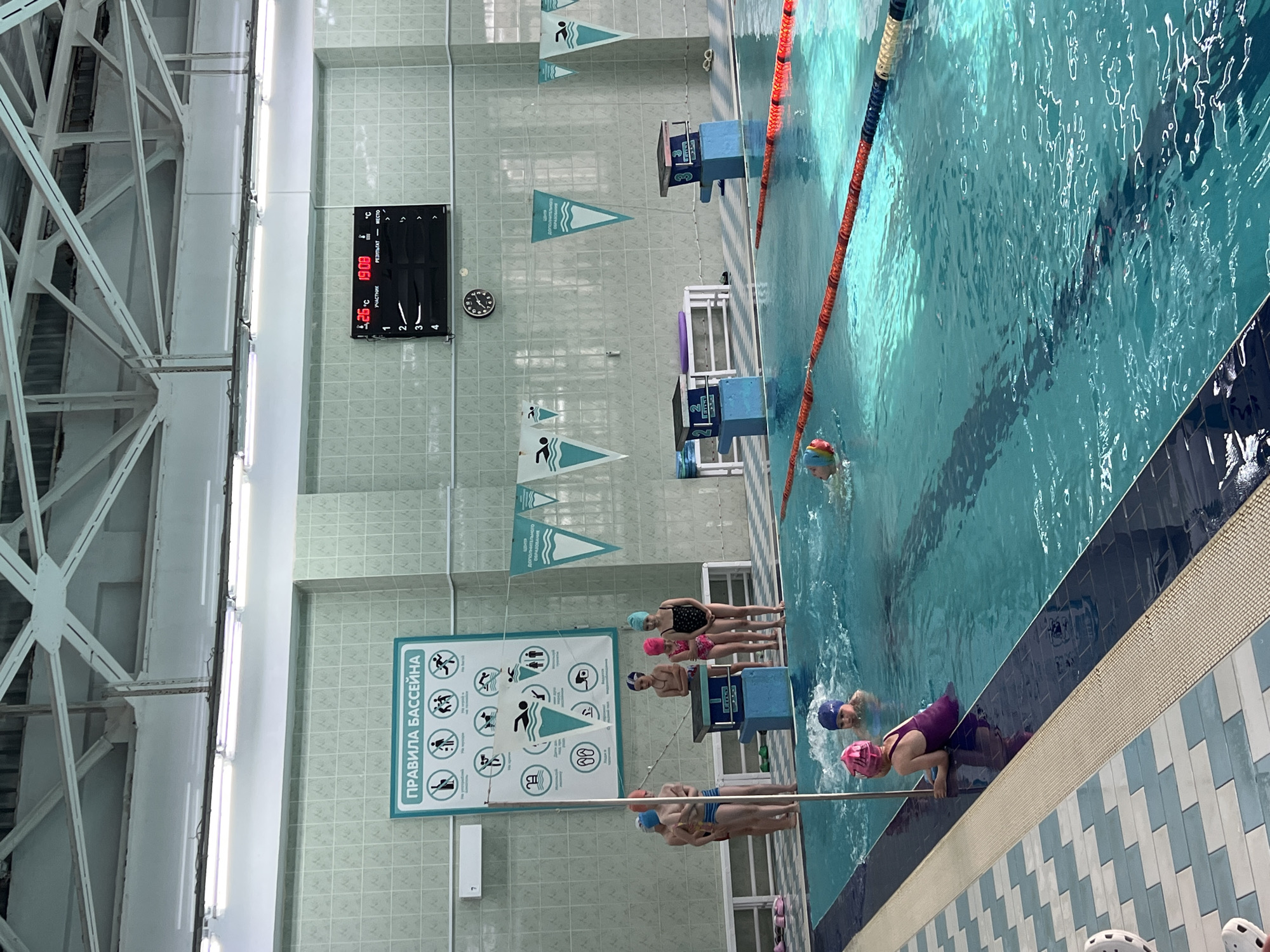 The image captures an indoor swimming pool, possibly a training facility or public pool, located in Марковский, Russia. The scene is set within a tiled room with a high, exposed metal framework ceiling and fluorescent lighting. A digital scoreboard displays information, including the time and temperature. Wall signage with pictograms illustrates swimming pool rules. Several children, wearing swim caps and swimsuits, are positioned at the pool's edge, some preparing to enter the water. An orange lane rope divides the pool. One person is swimming. Starting blocks with lane numbers are present. The pool water is clear and reflects the overhead light. The pool deck has tiled edging.