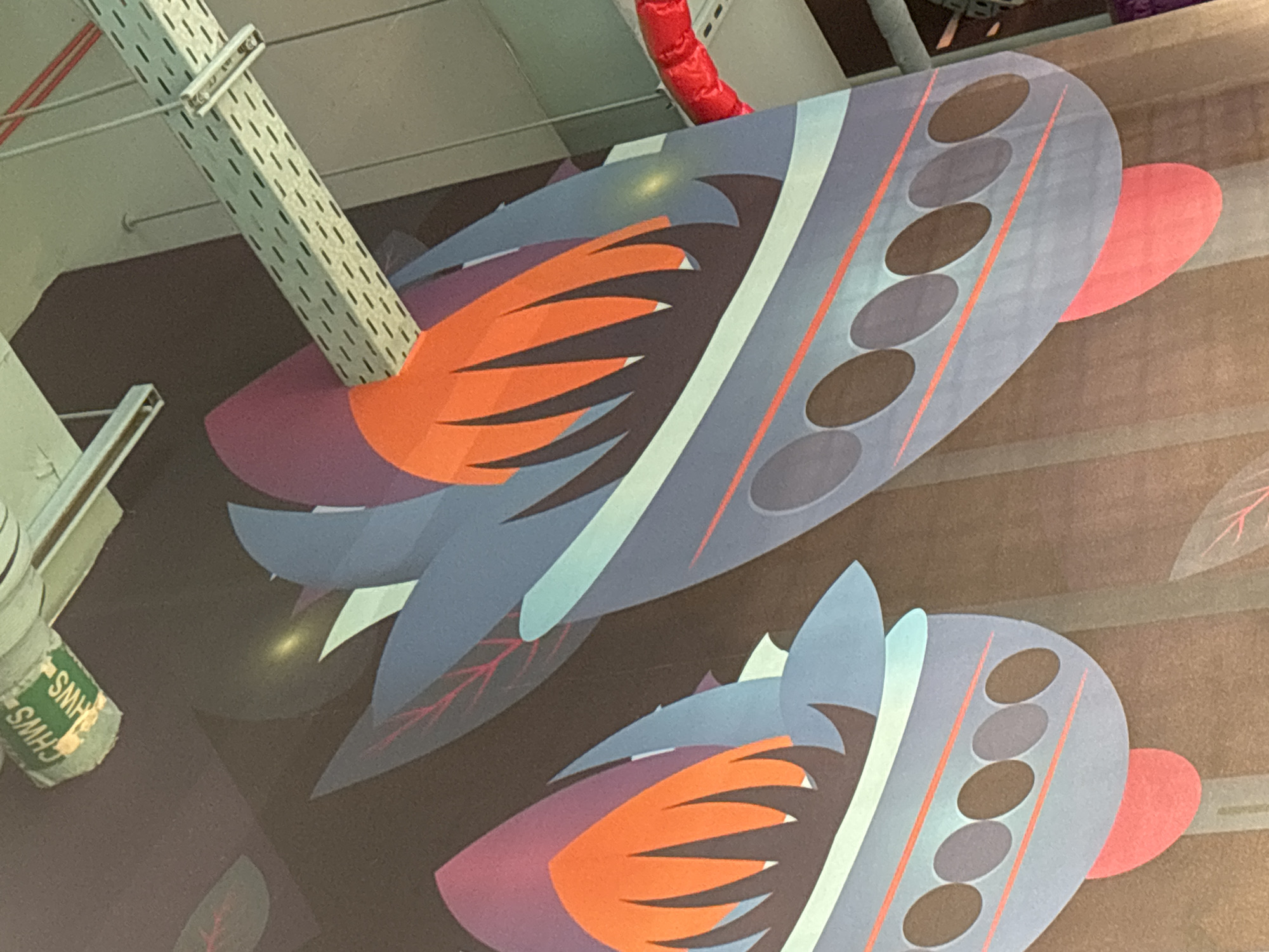 The image depicts a section of a large, colorful mural on a wall, possibly in Dubai, United Arab Emirates. The mural features abstract, stylized shapes in shades of blue, orange, purple, and pink, resembling fins or leaves. In the upper left corner, a small green sign is visible with white text that appears to read "CHWS HWS." Above the mural, metal conduits and lighting fixtures are present, suggesting an indoor setting, potentially a public space. The overall impression is of a vibrant, artistic decoration.