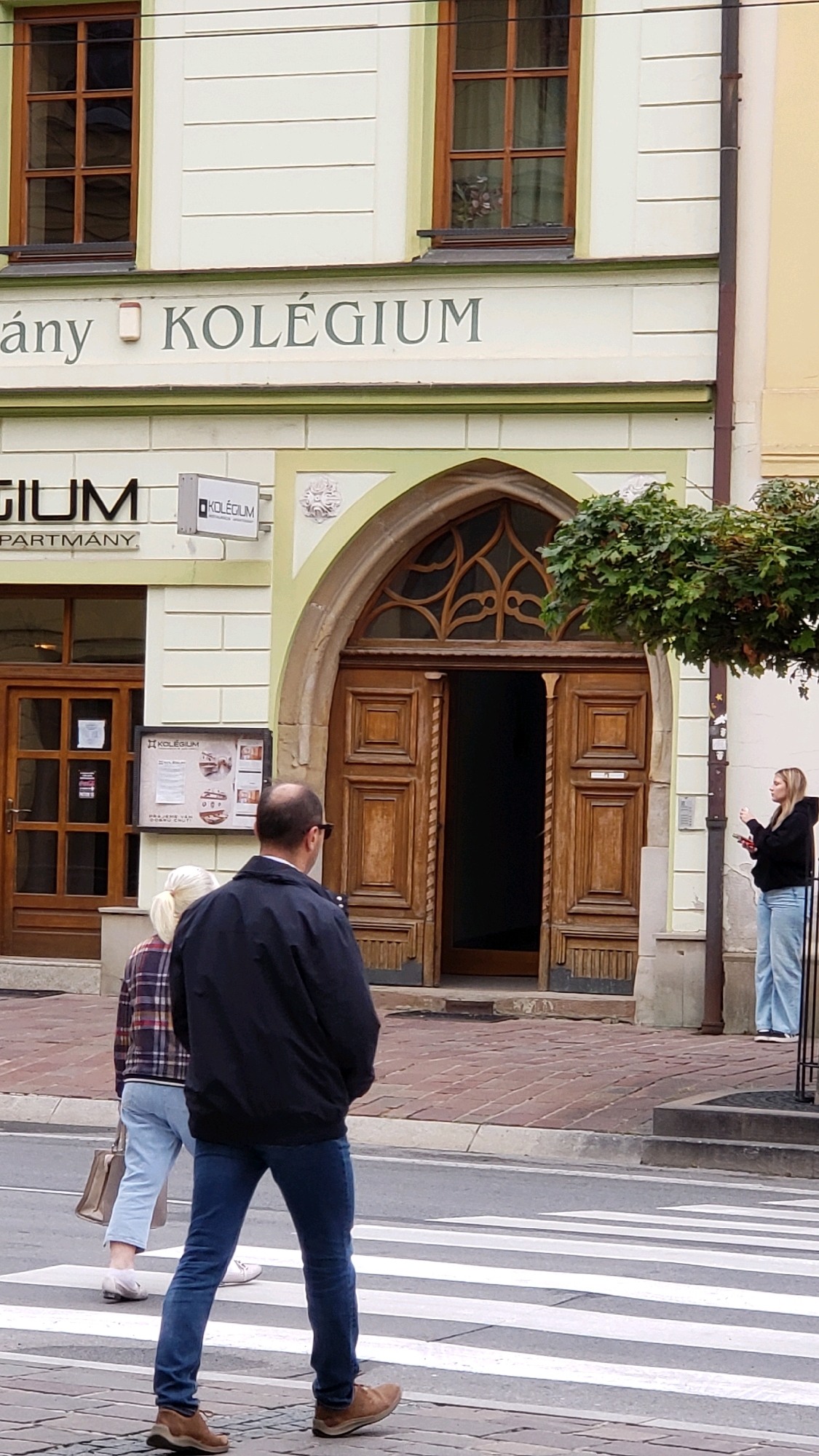 The image captures a street scene in front of a building with the sign "KOLÉGIUM" prominently displayed. The building appears to be located in Prešov, Slovakia, given the context provided.
In the foreground, two individuals are walking on a crosswalk. The person in the front is a man wearing a dark jacket and blue jeans, with brown shoes. The person behind him is a woman wearing a plaid shirt and light blue jeans, carrying a tan handbag. They are walking from left to right across the frame.
The building itself has a light-colored facade with dark wood-framed windows. There is a large, arched wooden doorway with intricate carvings. A sign for "KOLÉGIUM" is also visible on the building near the doorway. To the right of the doorway, a young woman is standing on the sidewalk, wearing a black hoodie and light blue jeans, holding a red object in her hand. She appears to be gesturing or talking.
The street in front of the building is paved with cobblestones and a pedestrian crosswalk is clearly marked. A tree with green foliage grows on the right side of the building. The overall environment suggests an urban setting, likely a historical part of the city.