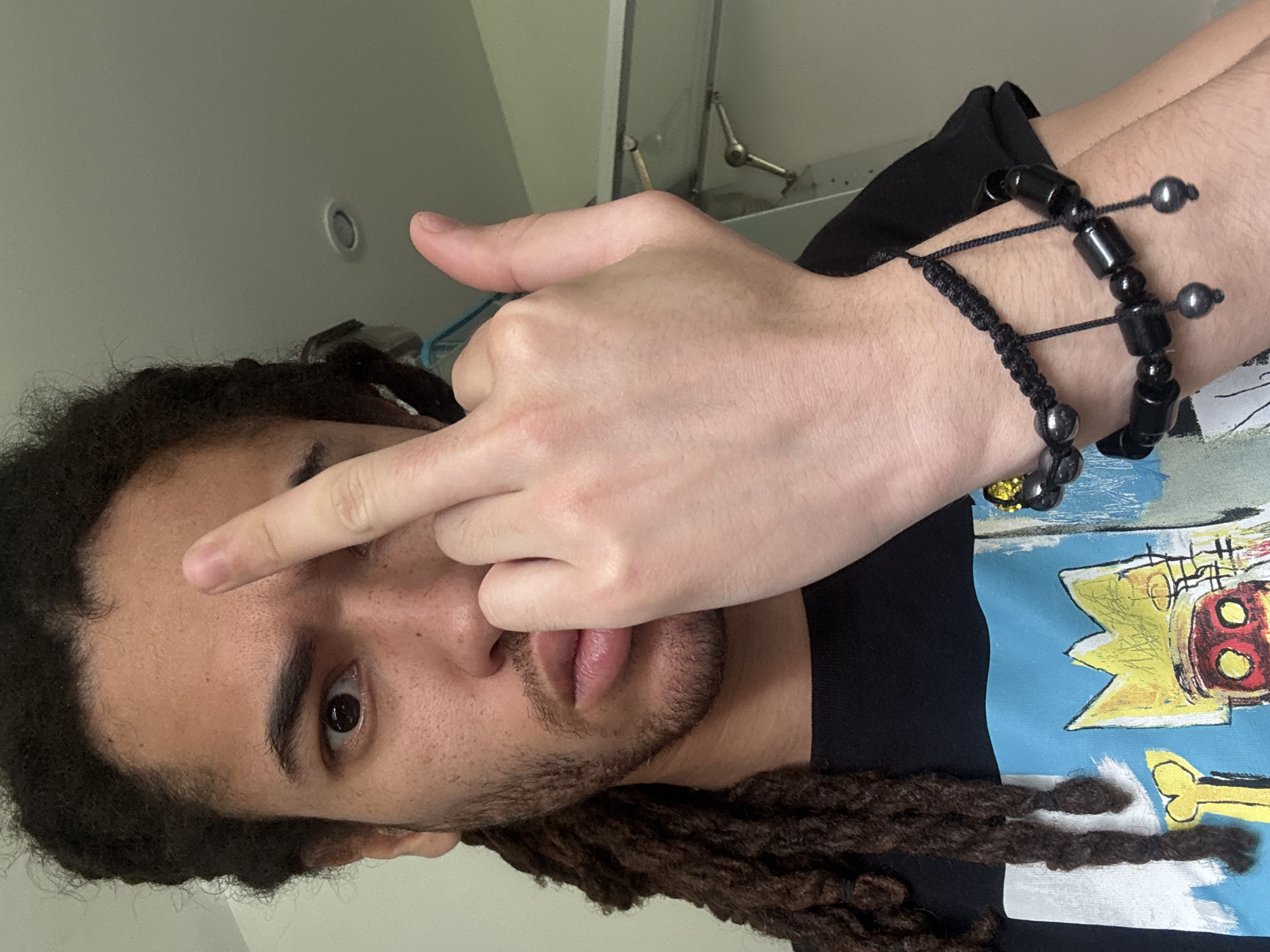 A man with dark hair styled in dreadlocks is giving the middle finger. He is wearing a black t-shirt with a colorful graphic print and two black bracelets on his right wrist. The background is a plain light-colored wall with a light fixture near the top left. The man has light brown skin, a trimmed beard, and brown eyes. There is no indication of location in Les Ulis, France.