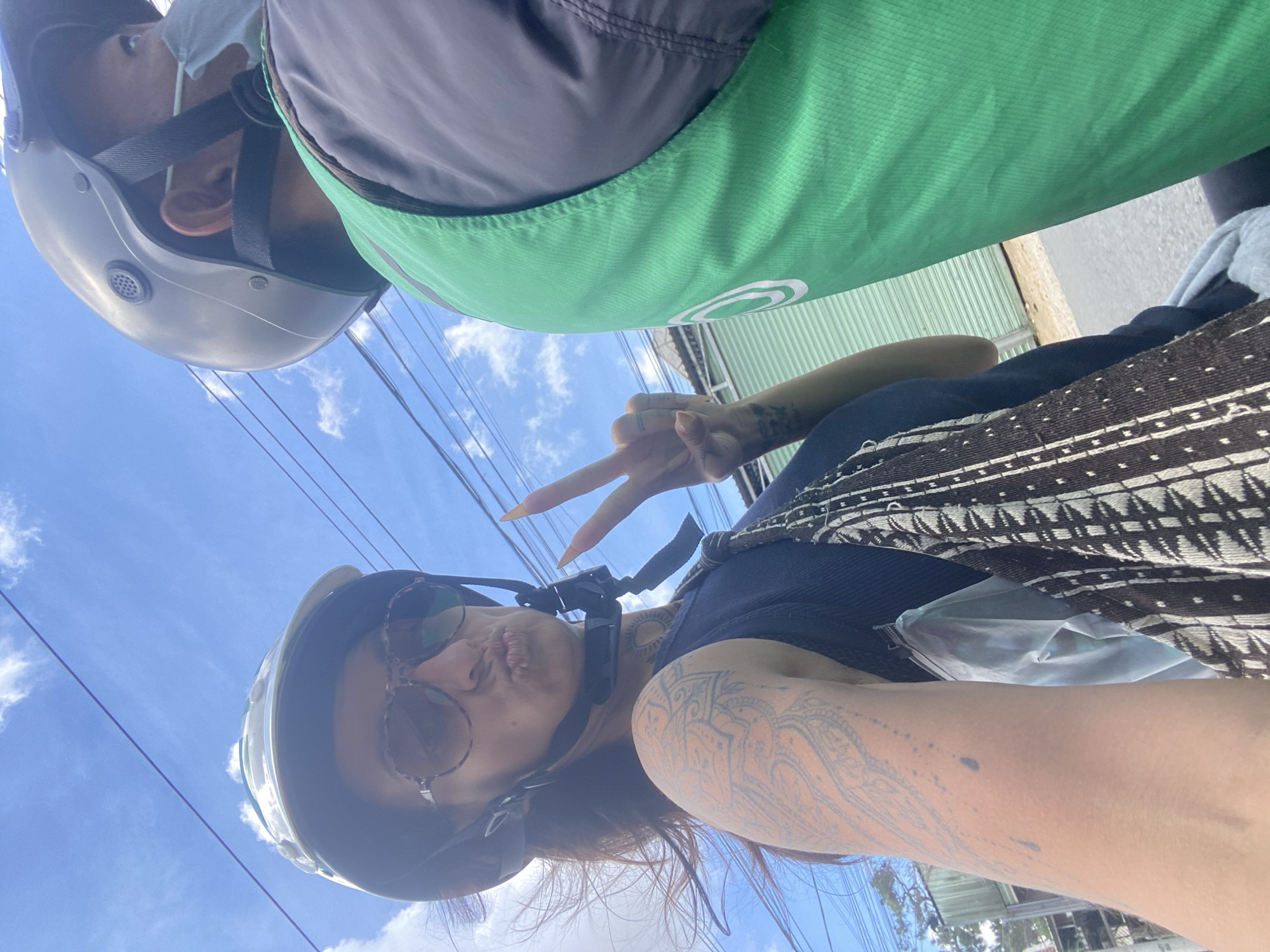 A medium shot captures two individuals on a motorcycle against a bright blue sky with wispy white clouds. The person in the foreground, a woman, wears a white helmet, large sunglasses, and a dark blue tank top. Her lips are pursed, and she holds up two fingers in a peace sign. She has extensive tattoos on her left arm, with a visible sun tattoo on her neck. A patterned bag is slung over her shoulder. The person behind her wears a dark helmet and a light blue face mask, along with a bright green vest. They are facing to the right, looking forward. The background shows power lines stretching across the sky and a building with a ribbed metal facade. The scene appears to be on a street in Da Lat, Vietnam, likely on a sunny day.