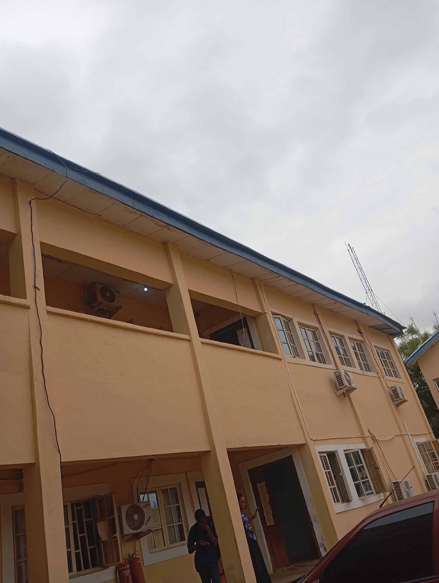 The image shows a light peach-colored building with a dark blue trim along the roofline. The building has multiple windows, some open and some closed. There are air conditioning units mounted on the exterior walls of the building. A tall antenna structure is visible against the cloudy sky. The overall scene suggests an urban environment. The specific location is the Ministry of Information and Orientation building in Jalingo, Nigeria. The image appears to have been taken during the day under overcast skies.