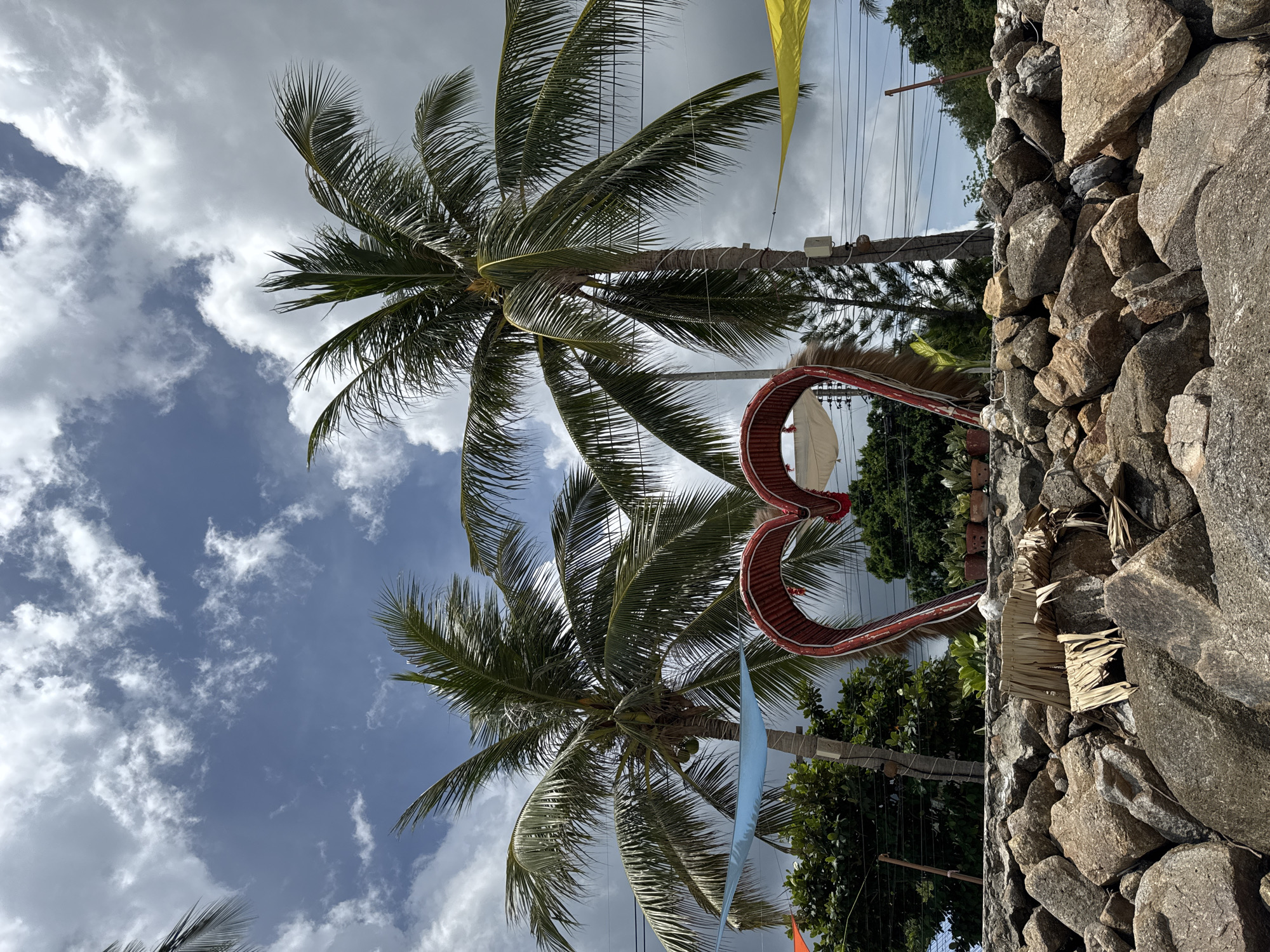 The image shows a view from below, looking up at two coconut palm trees set against a backdrop of a partly cloudy sky. In the foreground, to the right, is a stone wall, composed of large, irregularly shaped rocks. Adjacent to the wall, there is a decorative heart-shaped structure made from a red material. The palm trees are adorned with decorations. The photograph appears to be taken in Baan Chaloklum, Thailand.