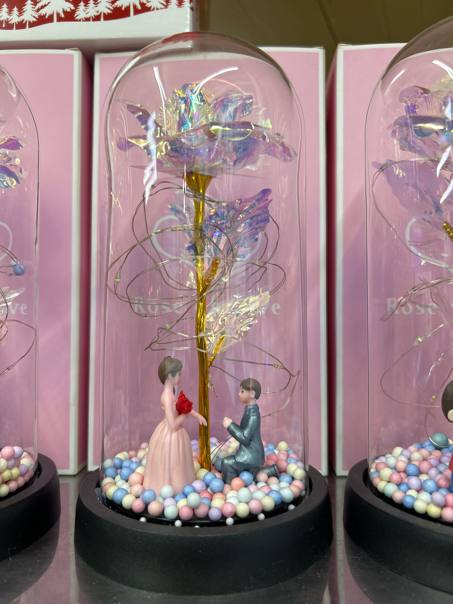 The image shows a close-up of three decorative glass domes, each containing a rose and miniature figurines. The rose in the central dome is iridescent and appears to be made of foil or plastic, with delicate LED lights wrapped around its stem. Inside this dome, a male figurine in a suit is on one knee, holding a red rose to a female figurine in a pink dress. Both figurines are surrounded by a bed of small, colorful, bead-like decorations. The domes are displayed in front of pink boxes with what appears to be white or red Christmas tree graphics on the top box. The surface beneath the domes is reflective, suggesting a shelf or table. The scene appears to be indoors, possibly in a retail setting. There is no indication of the location being in Buenos Aires, Argentina.