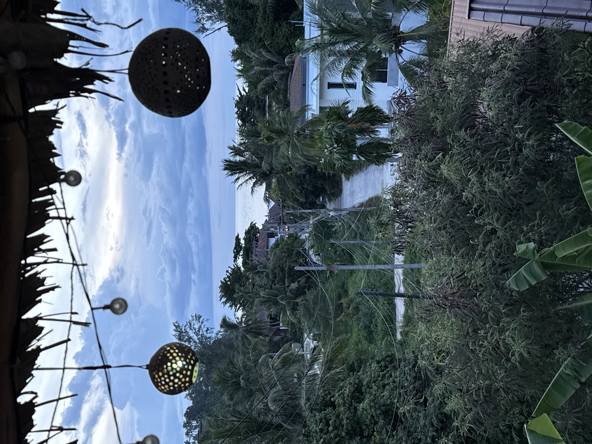 The image depicts an elevated view of a tropical coastal landscape in ตำบลเกาะพะงัน, Thailand, observed during the late afternoon or early evening. In the immediate foreground, a thatched roof structure is partially visible overhead, from which a string of lights is suspended. Two decorative spherical lanterns hang from these lights; the one on the left emits a warm glow through its perforations, while the one on the right is silhouetted against the sky.
The upper midground is dominated by an overcast sky with blue and white clouds. Below the sky, a calm expanse of ocean stretches horizontally across the scene.
The terrestrial midground and background feature dense, lush tropical vegetation, including numerous palm trees and various broadleaf trees. A narrow, winding paved road traverses this vegetation, appearing to lead towards the ocean. Multiple utility poles are erected along the road and throughout the landscape, supporting a complex network of overhead power lines that crisscross the view.
Several buildings are interspersed among the foliage. A distinct modern white building with a flat roof and prominent dark-framed vertical windows is visible on the right side of the image. Other structures with pitched roofs, including one with reddish-brown tiles and another with darker grey tiles, are partially discernible amidst the greenery in the background. No people or animals are present in the scene.