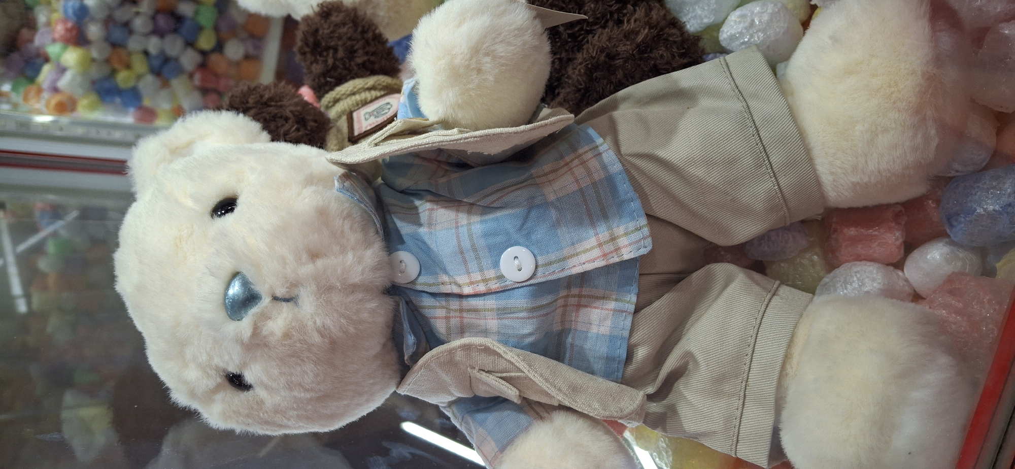 The image shows a close-up of a cream-colored teddy bear dressed in a blue plaid shirt, a light brown vest, and tan shorts. The teddy bear is sitting amongst colorful, bubble-wrapped objects that appear to be candy or small toys. In the background, more toys are visible in a display case, suggesting a store or arcade. The location is stated to be in the city of Telmana, Russia.