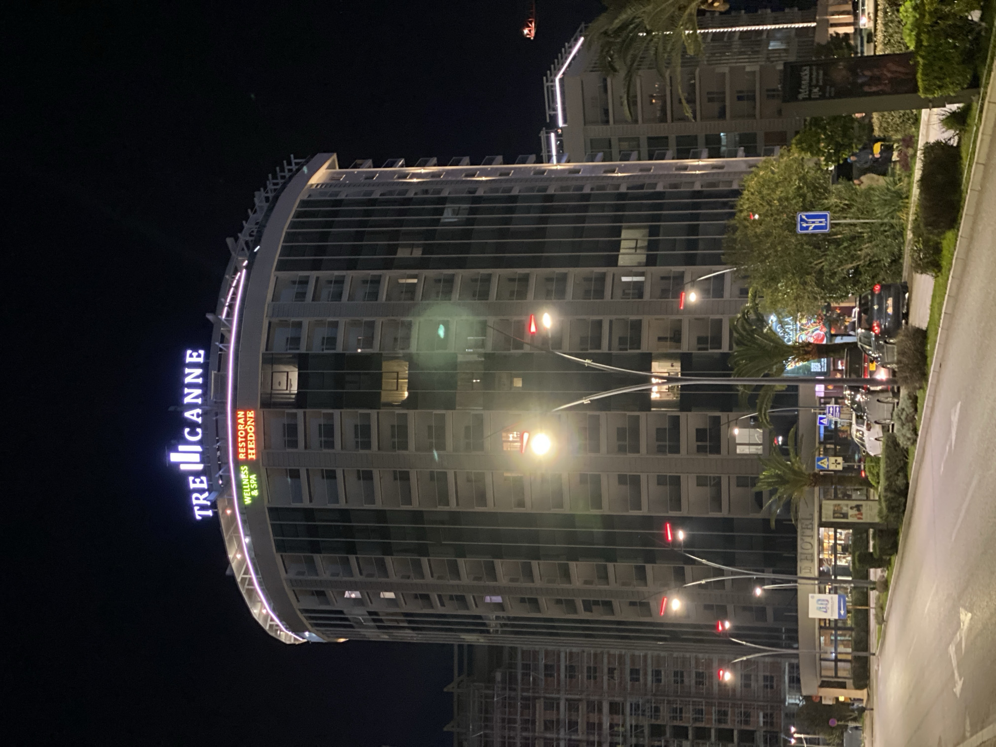 The image displays a nighttime street scene in Budva, Montenegro, dominated by the illuminated "Tre Canne" building.
The central subject is a tall, modern, cylindrical building, "Tre Canne," with multiple stories of balconies and windows, some internally lit. The building's rooftop features prominent white/blue neon signage for "TRE CANNE." Below this, on a lower level, additional illuminated signs read "WELLNESS & SPA" in green and "RESTORAN HEDONE" in red.
In the foreground, a multi-lane asphalt road with visible white lane markings extends from the bottom of the frame. A median strip separates the road lanes, densely planted with trimmed bushes, small palm trees, and other green foliage. Multiple ornate streetlights with bright, warm-toned lamps and smaller red horizontal lights are spaced along this median, illuminating the scene.
Several vehicles are present on the road and alongside the median. A dark hatchback car, possibly a Volkswagen Golf, is visible near the right side of the median, with other cars parked or moving further down the street. Various street signs are visible on the median, including a blue square sign depicting diverging arrows and another blue sign indicating pedestrian crossing. A dark, vertical informational or advertising board stands on the right side of the median.
Multiple individuals are visible on the sidewalk and median area. A group of approximately three to four people is discernible on the right side of the median, near the dark vertical sign. One individual in this group is wearing a blue hooded garment, and another a lighter-colored jacket. Another lone figure is visible on the sidewalk near a ground-level entrance to the "Tre Canne" building, where a sign for "HOTEL" is also present. No specific actions or interactions are clearly discernible beyond standing or walking.
In the background, to the left of the "Tre Canne" building, another tall structure is visible, partially covered with scaffolding, indicating ongoing construction. To the right, additional illuminated buildings and palm trees are present. A red construction crane boom with lights is visible in the upper-right distance against the dark night sky.